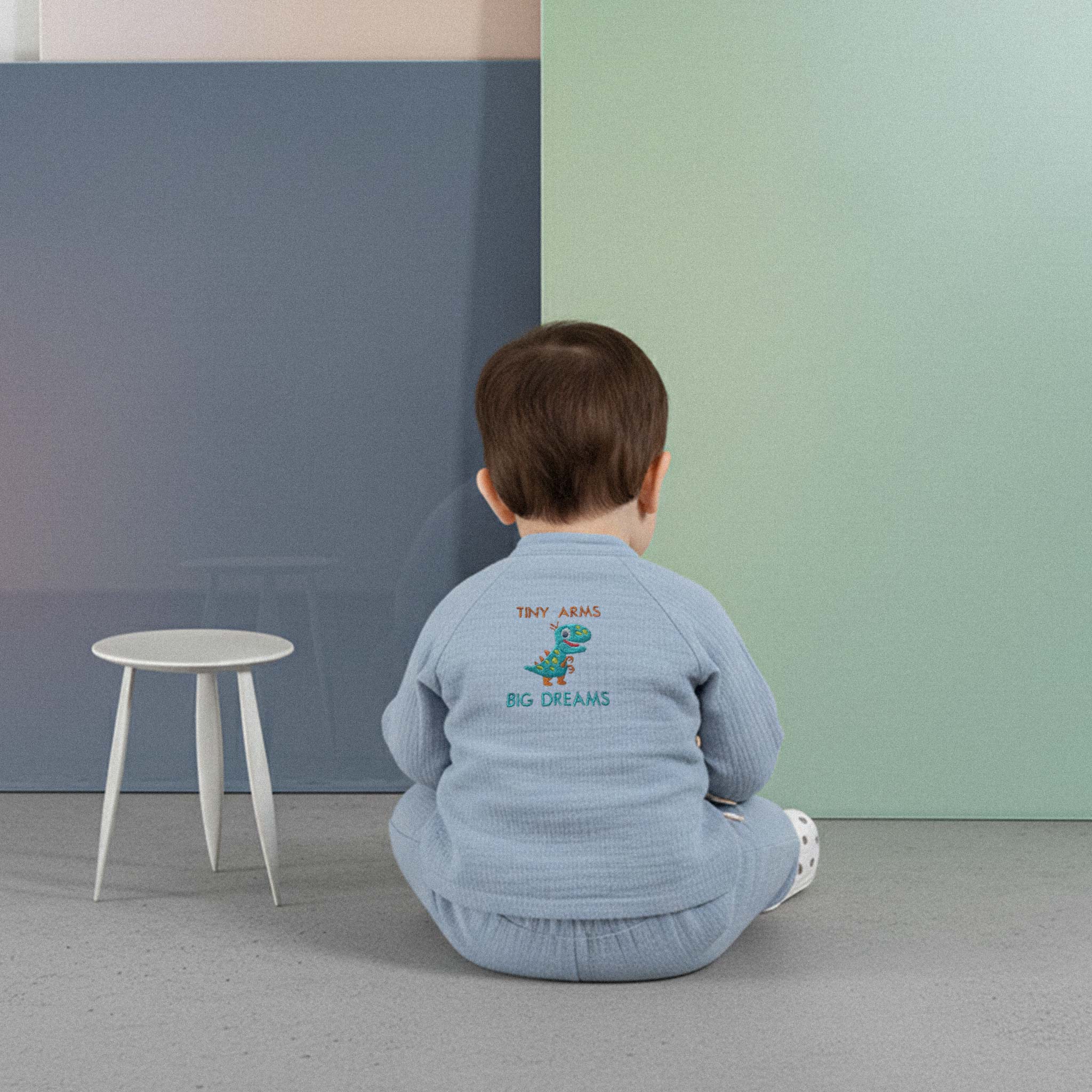 Üzerinde indigo mavisi renkli, 'Tiny Arms Big Dreams' dinozor baskılı müslin kimono takımı bulunan sevimli bebek, renkli küplerin önünde yerde otururken kollarını açarak neşeyle gülümsüyor.