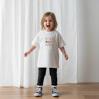 Siyah tayt ve ekru renkli, 'Too Good for Broccoli' nakışlı reglan kollu oversize bebek tişörtü giyen küçük kız çocuk, neşeyle kollarını açarak poz veriyor.