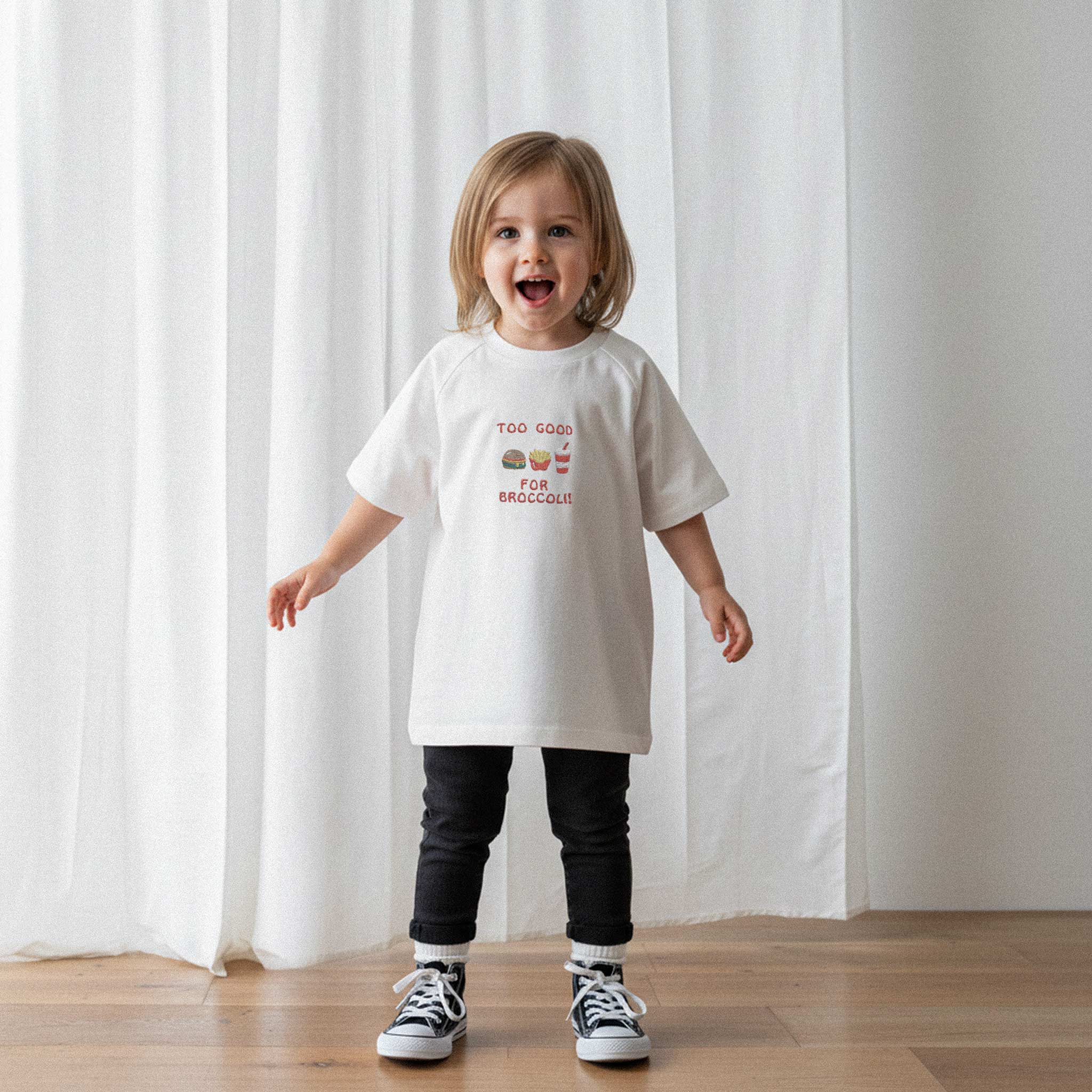 Siyah tayt ve ekru renkli, 'Too Good for Broccoli' nakışlı reglan kollu oversize bebek tişörtü giyen küçük kız çocuk, neşeyle kollarını açarak poz veriyor.