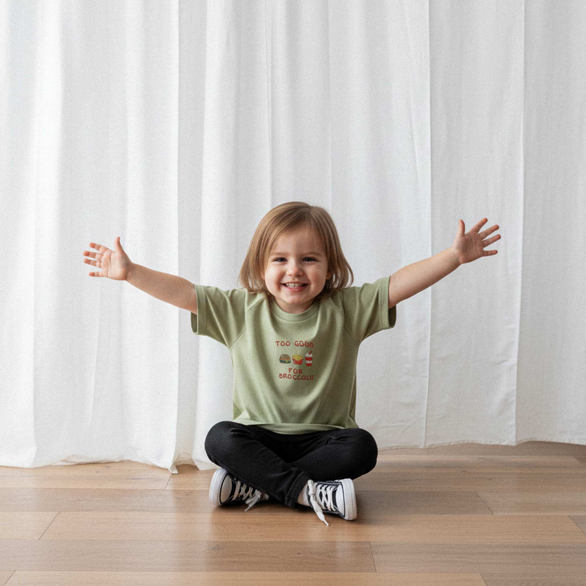 Siyah pantolon ve haki yeşili, 'Too Good for Broccoli' nakışlı reglan kollu oversize bebek tişörtü giyen küçük kız çocuk, yerde otururken kollarını açarak neşeyle gülümsüyor.