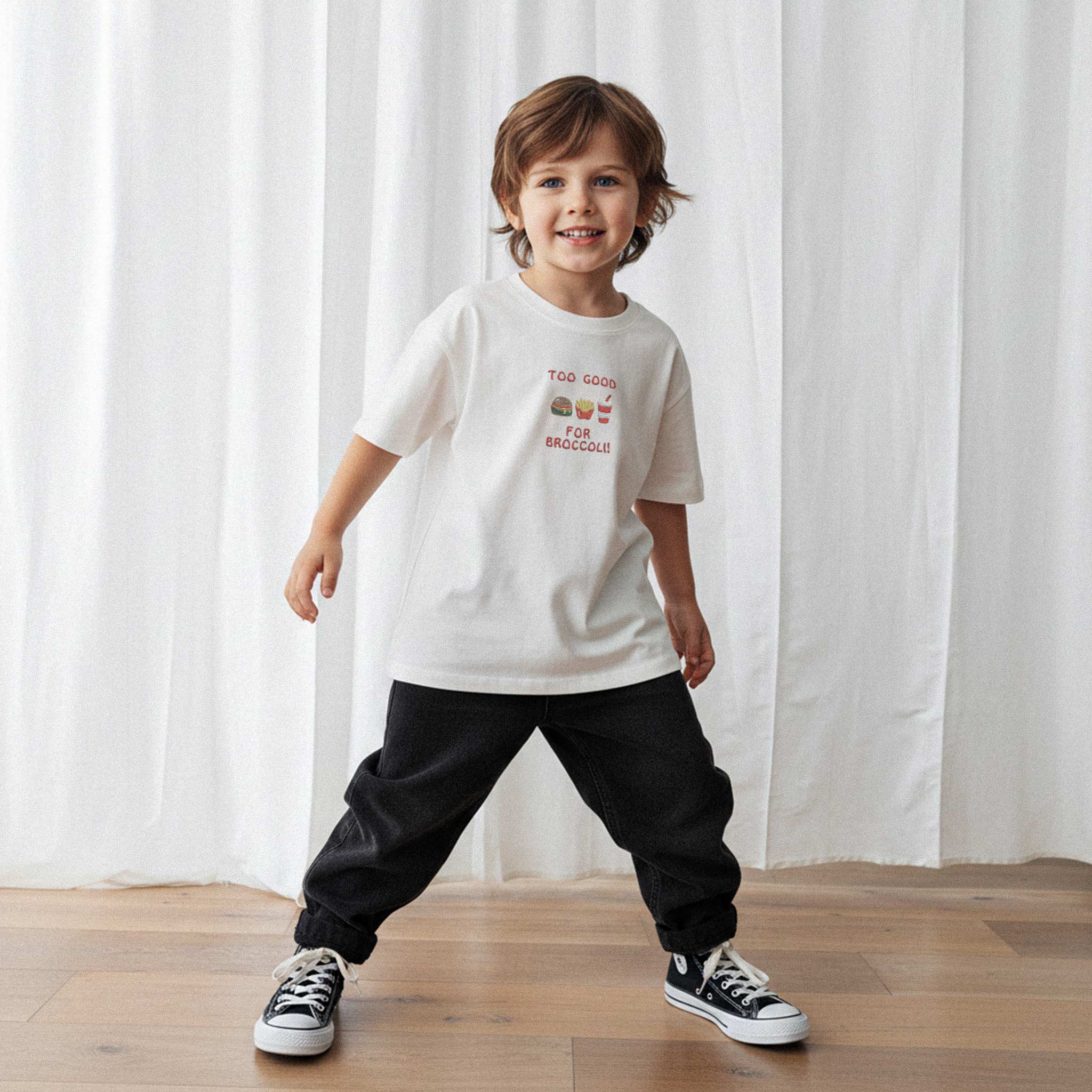 Siyah pantolon ve ekru renkli, 'Too Good for Broccoli' nakışlı oversize tişört giyen erkek çocuk, gülümseyerek kameraya bakıyor.