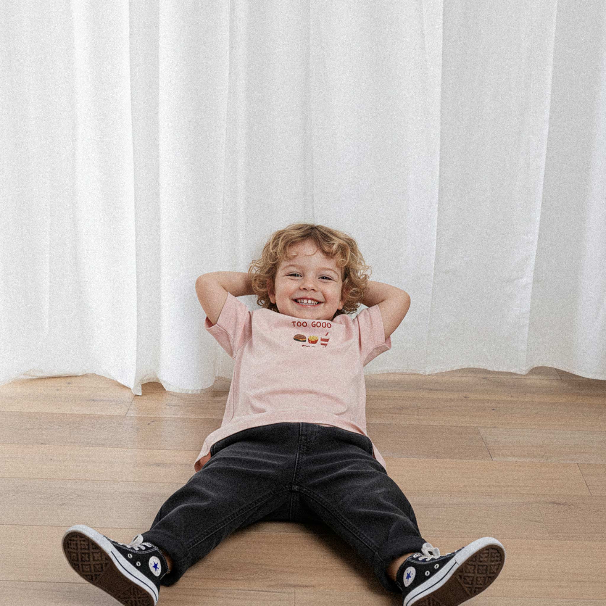 Siyah pantolon ve gül kurusu, 'Too Good for Broccoli' nakışlı oversize tişört giyen kıvırcık saçlı küçük erkek çocuk, yerde yatarken elleri ensesinde neşeyle gülümsüyor.