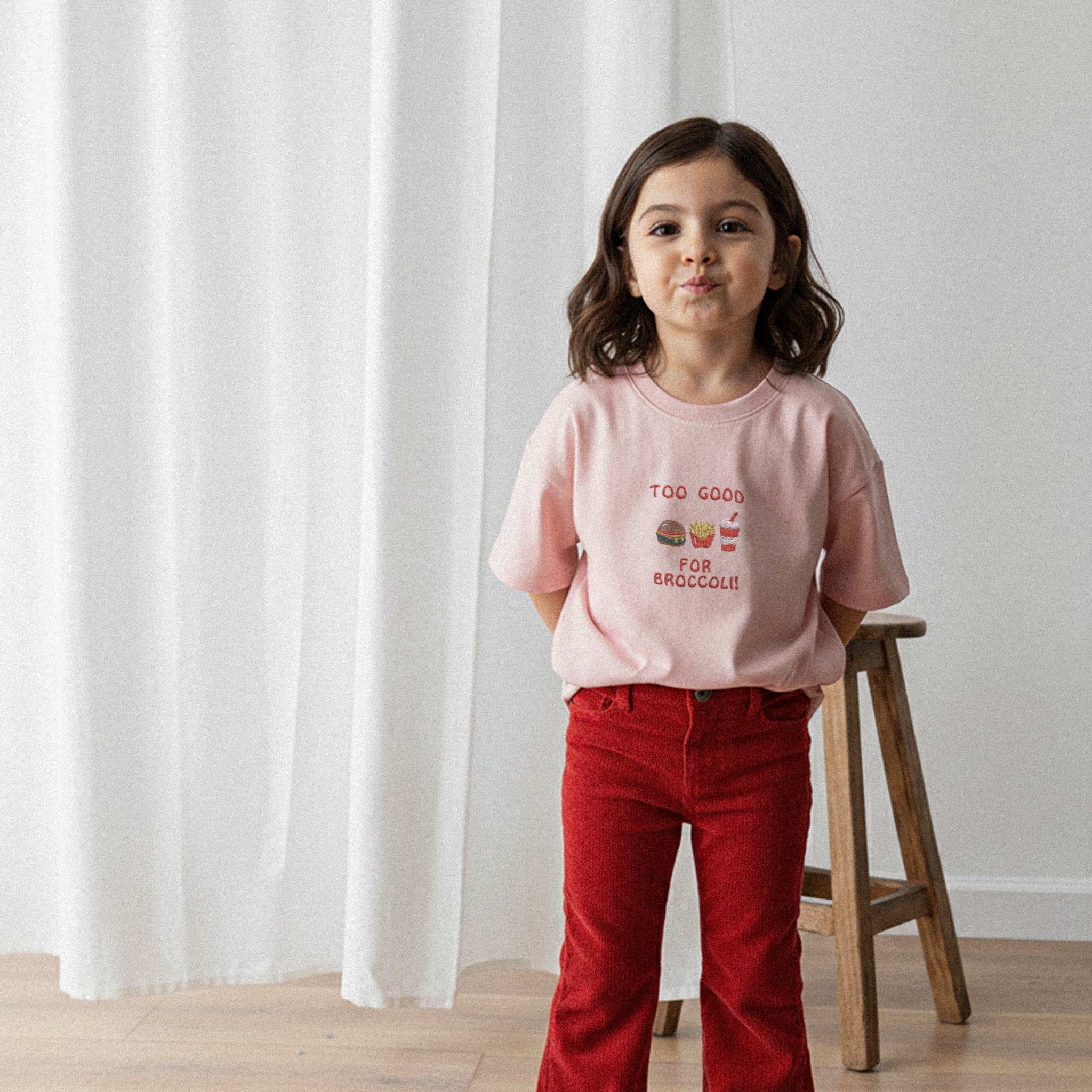 Kırmızı kadife pantolon ve gül kurusu, 'Too Good for Broccoli' nakışlı oversize tişört giyen kız çocuk, elleri arkasında sevimli bir poz veriyor.