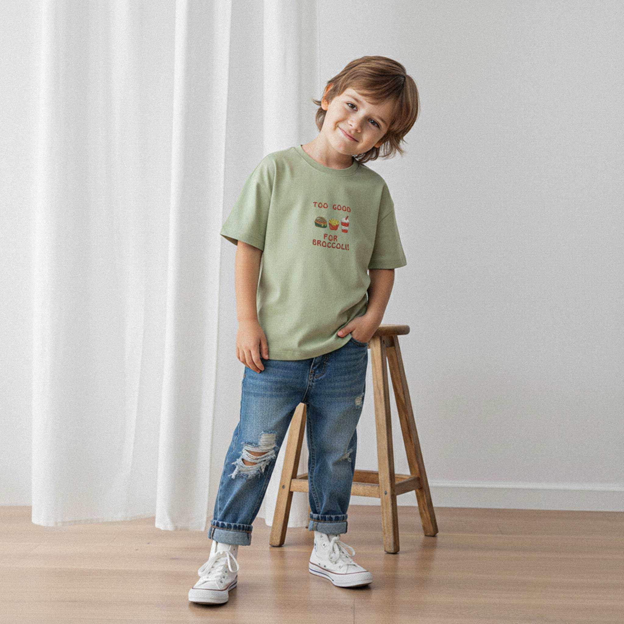 Yırtık jean pantolon ve haki yeşili, 'Too Good for Broccoli' nakışlı oversize tişört giyen erkek çocuk, bir eli cebinde gülümseyerek poz veriyor.