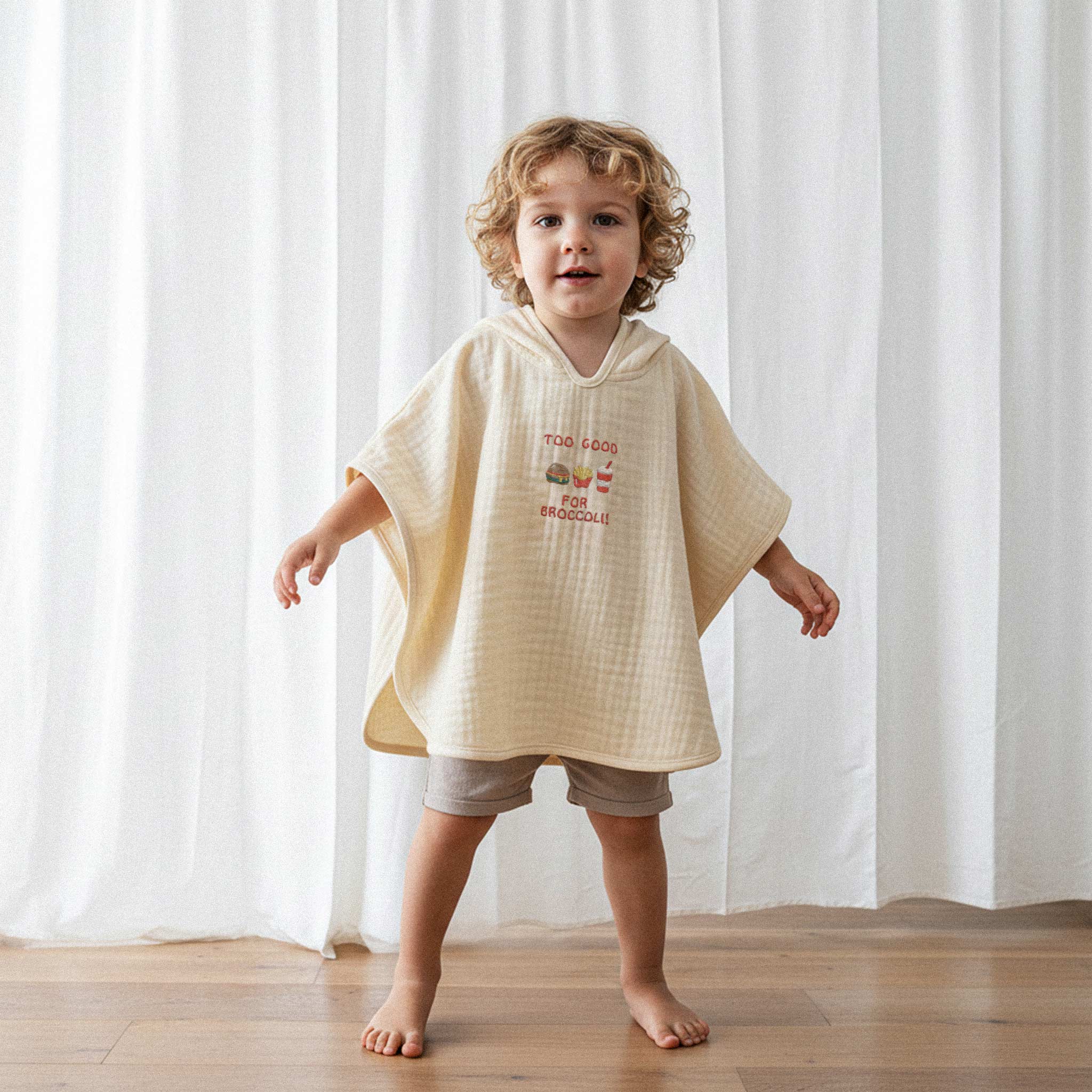 Bej şort ve ekru renkli, 'Too Good for Broccoli' nakışlı kapüşonlu müslin panço giyen kıvırcık saçlı küçük erkek çocuk, kollarını açarak poz veriyor.