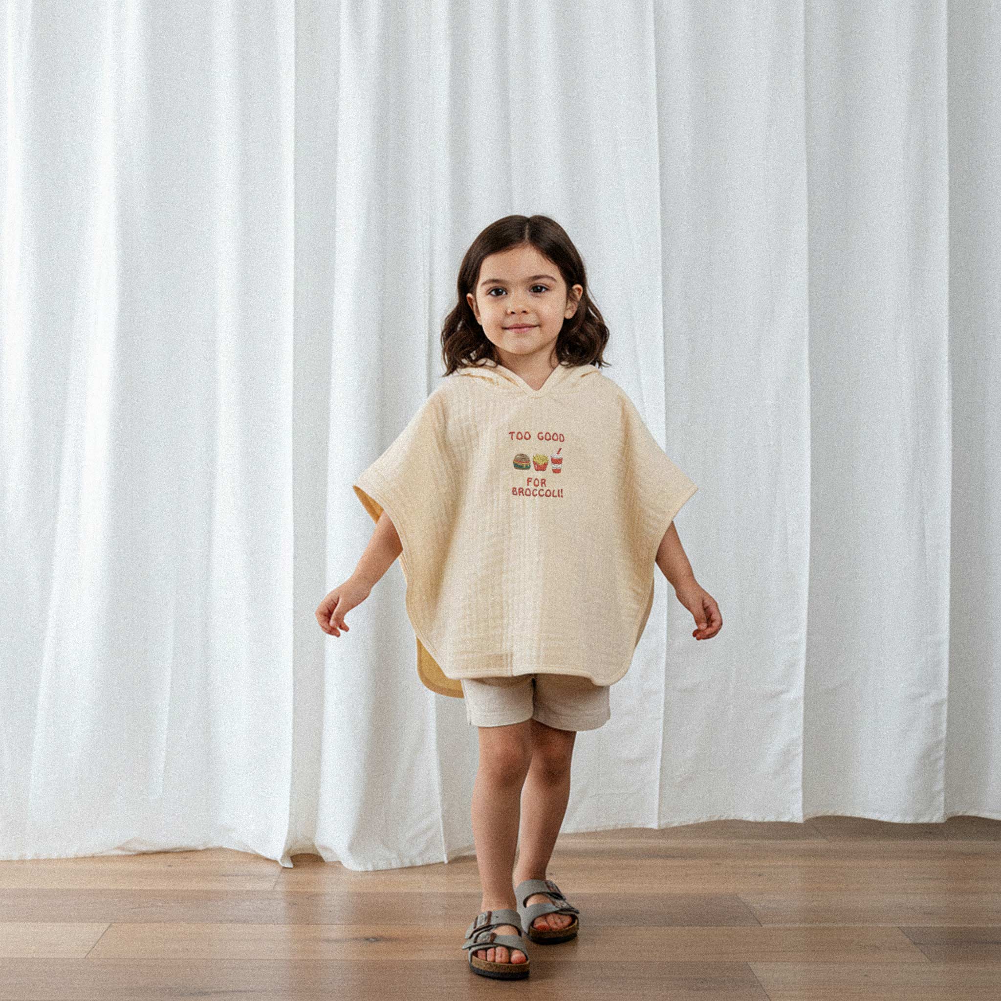 Bej şort ve sandalet giyen kız çocuk, üzerinde ekru renkli, 'Too Good for Broccoli' nakışlı kapüşonlu müslin panço ile gülümseyerek yürüyor.