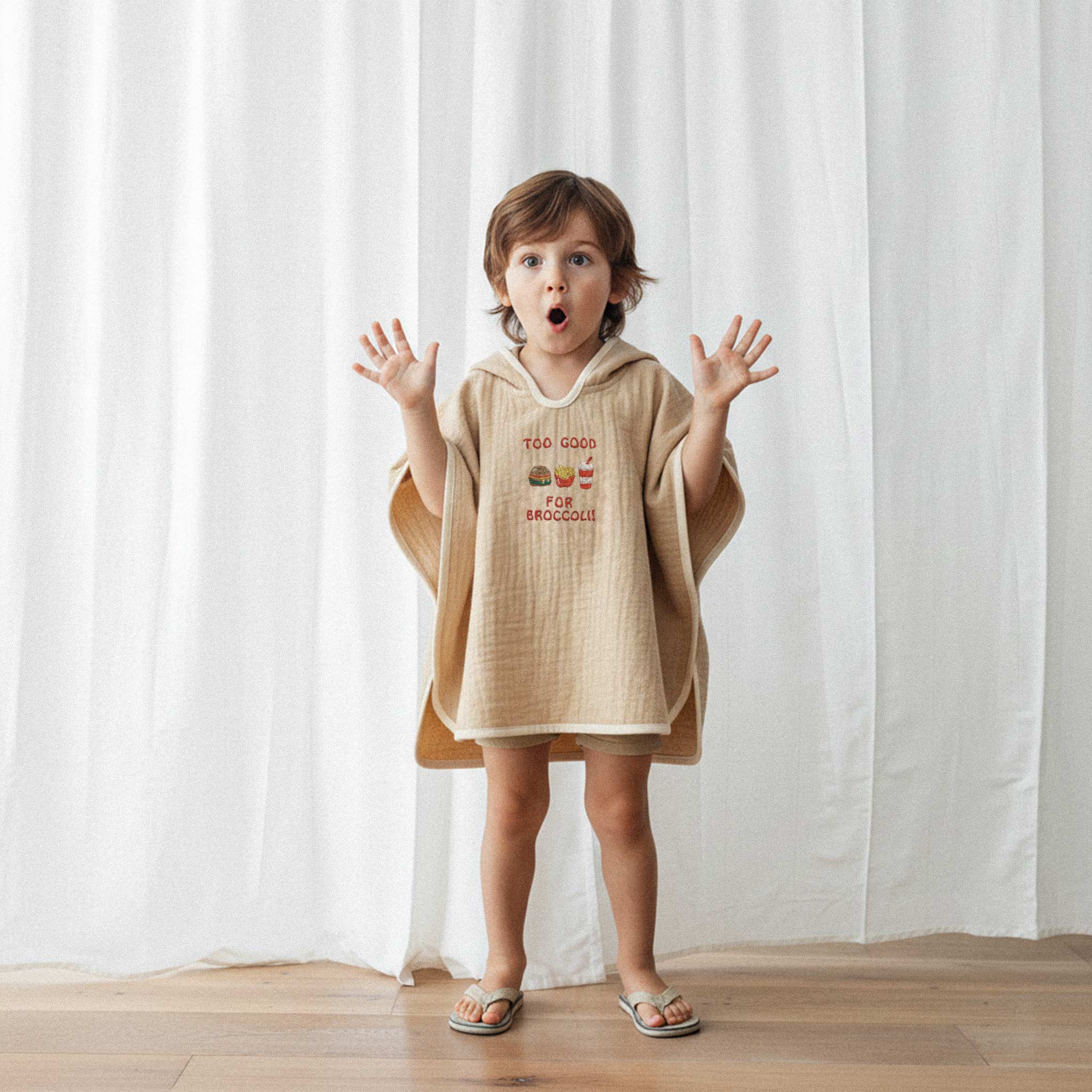 Bej şort ve somon rengi, 'Too Good for Broccoli' nakışlı kapüşonlu müslin panço giyen erkek çocuk, şaşkın bir surat ifadesiyle ellerini kaldırarak poz veriyor.