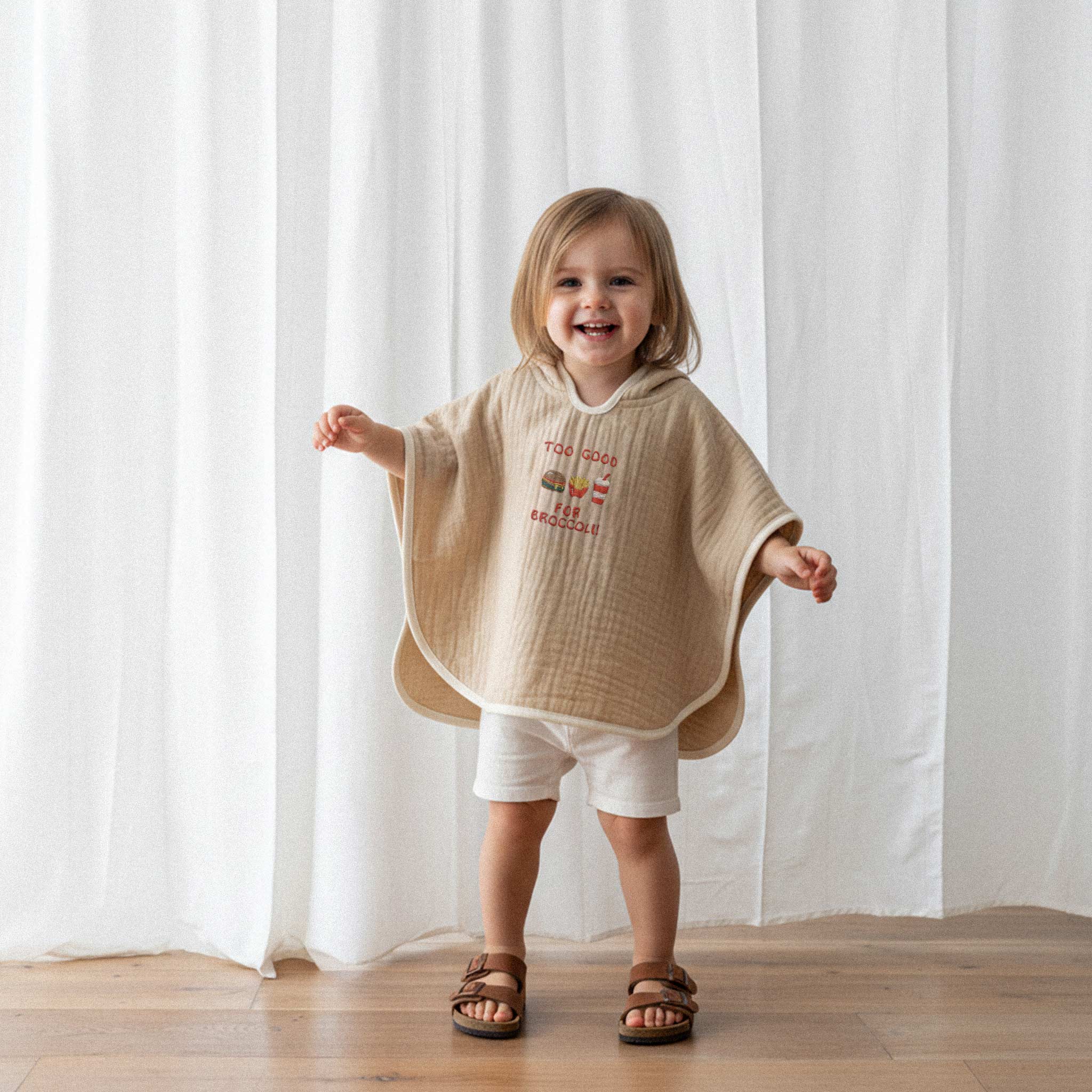 Beyaz şort ve somon rengi, 'Too Good for Broccoli' nakışlı kapüşonlu müslin panço giyen küçük kız çocuk, kollarını açarak neşeyle gülümsüyor.