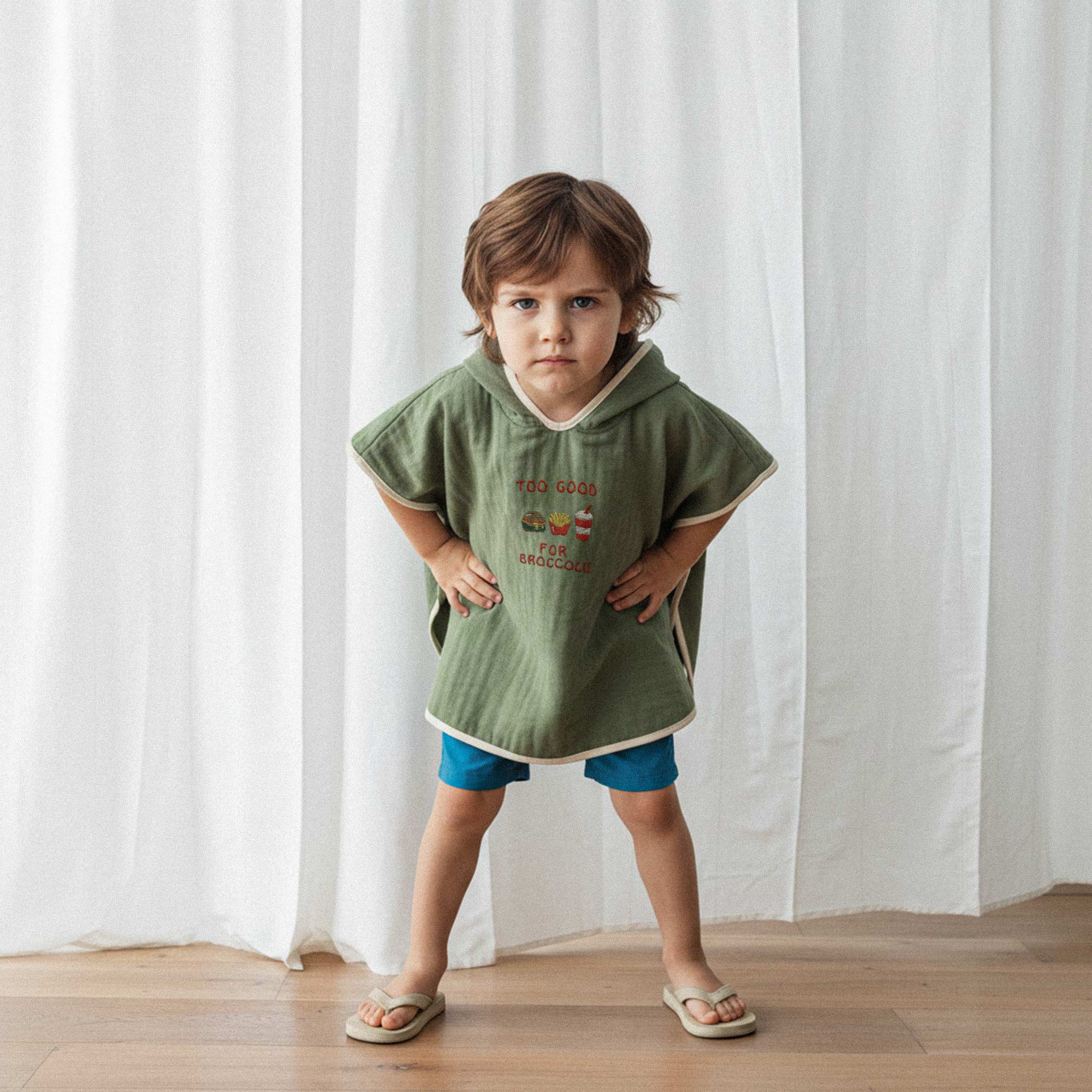 Mavi şort ve haki yeşili, 'Too Good for Broccoli' nakışlı kapüşonlu müslin panço giyen erkek çocuk, elleri belinde havalı bir şekilde poz veriyor.