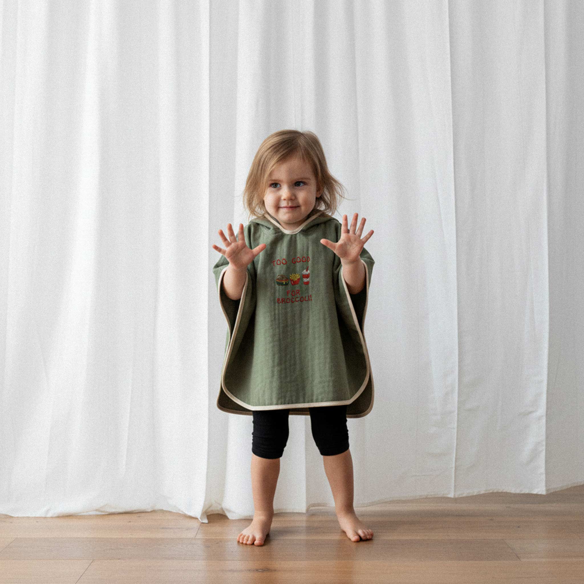 Siyah tayt ve haki yeşili, 'Too Good for Broccoli' nakışlı kapüşonlu müslin panço giyen küçük kız çocuk, ellerini kaldırarak sevimli bir şekilde poz veriyor.
