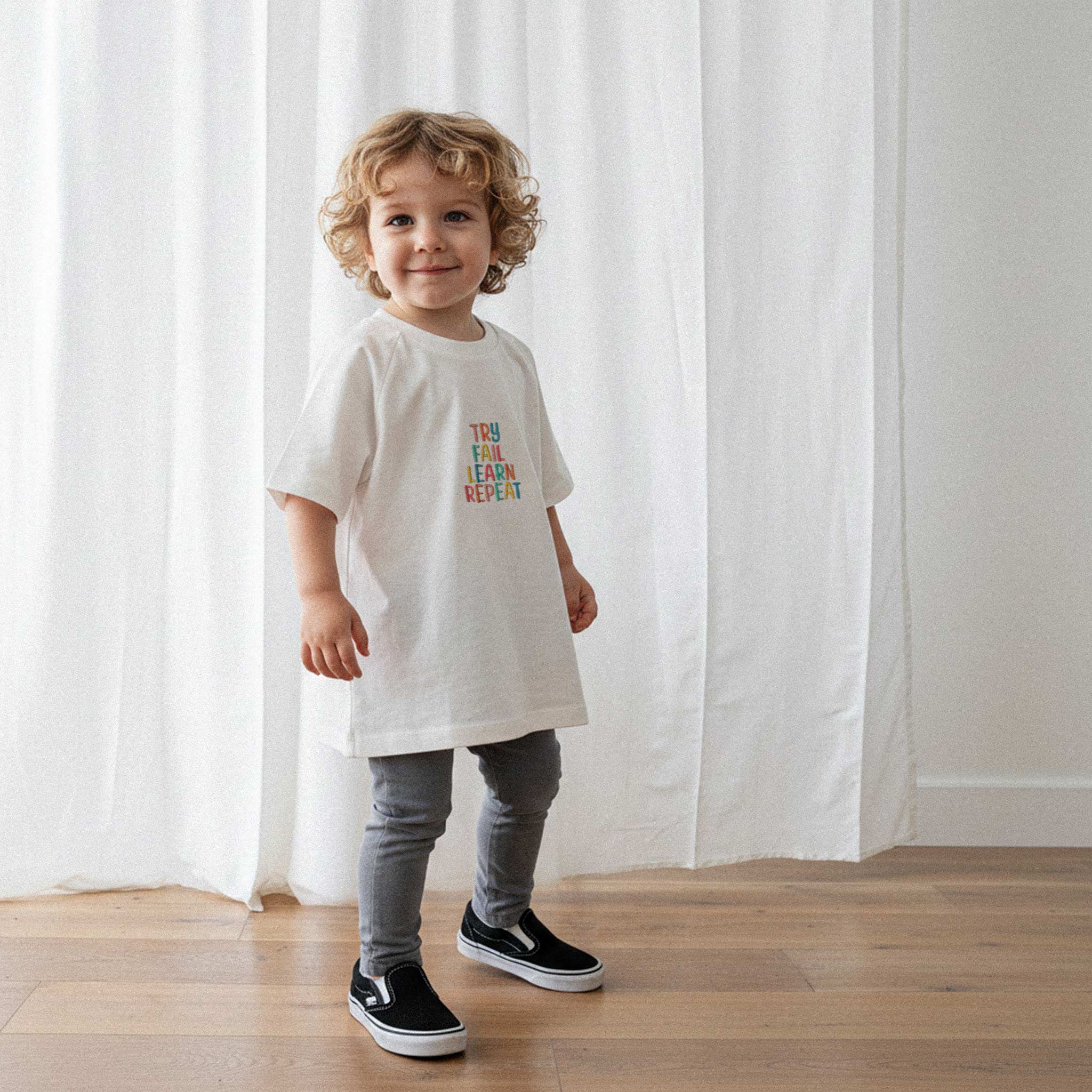 Gri tayt ve ekru renkli, 'Try Fail Learn Repeat' nakışlı reglan kollu oversize bebek tişörtü giyen kıvırcık saçlı küçük erkek çocuk, sevimli bir şekilde gülümseyerek poz veriyor.
