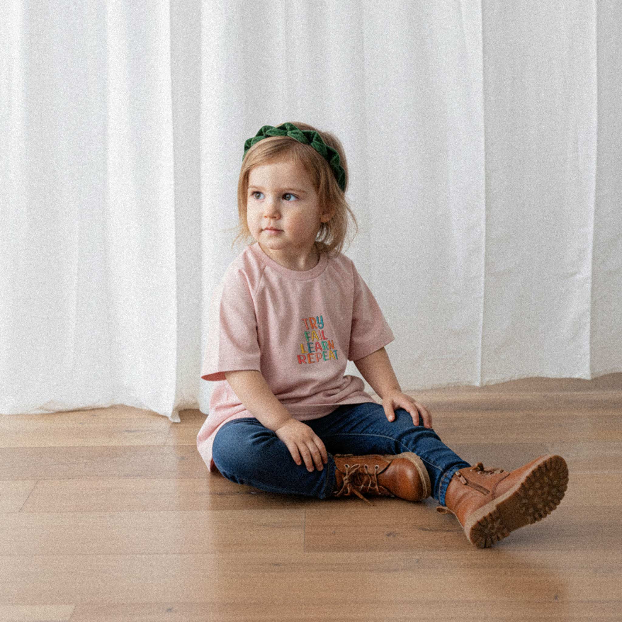 Jean pantolon ve gül kurusu renkli, 'Try Fail Learn Repeat' nakışlı reglan kollu oversize bebek tişörtü giyen küçük kız çocuk, yerde otururken düşünceli bir şekilde yana bakıyor.