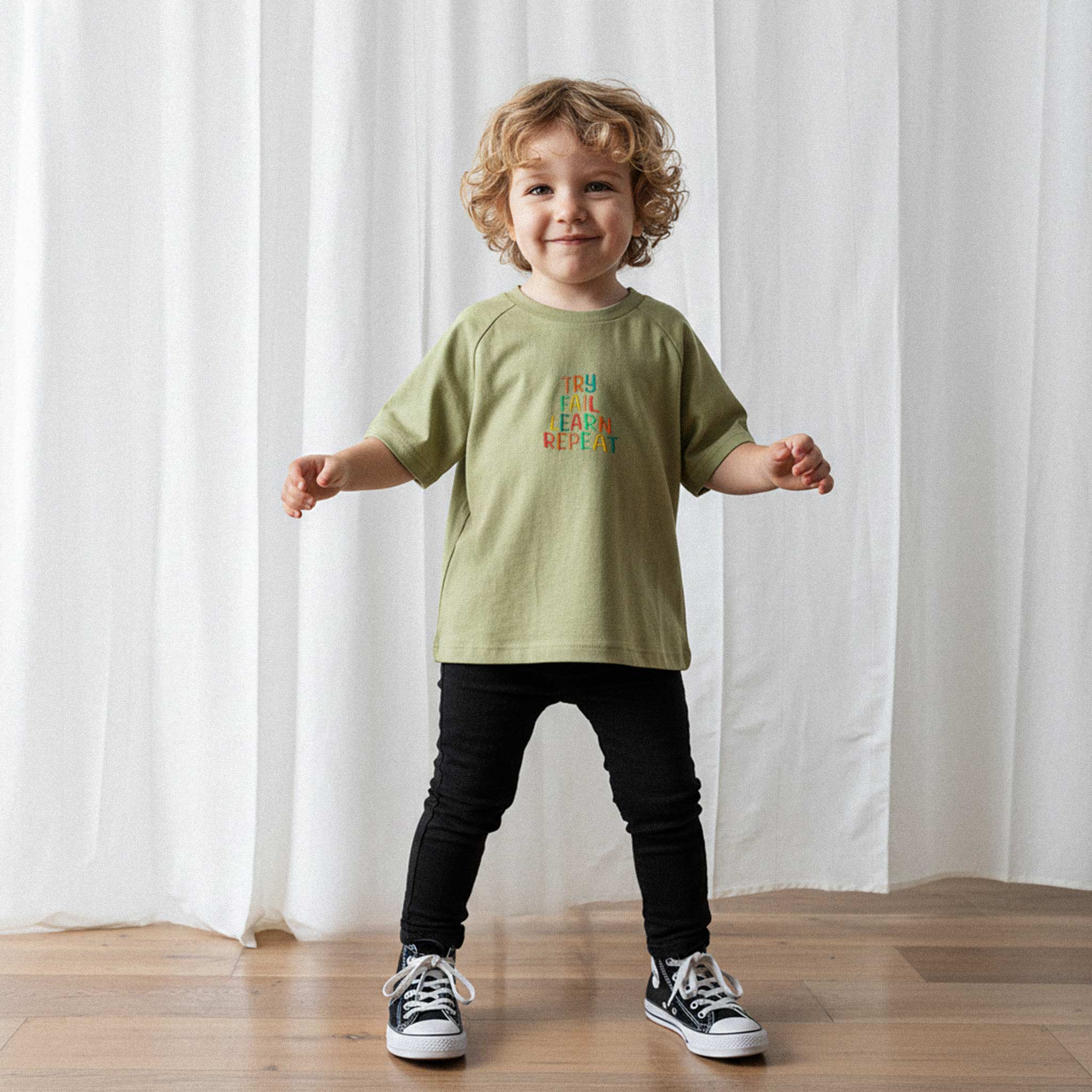 Siyah pantolon ve haki yeşili renkli, 'Try Fail Learn Repeat' nakışlı reglan kollu oversize bebek tişörtü giyen kıvırcık saçlı küçük erkek çocuk, sevimli bir şekilde gülümseyerek poz veriyor.