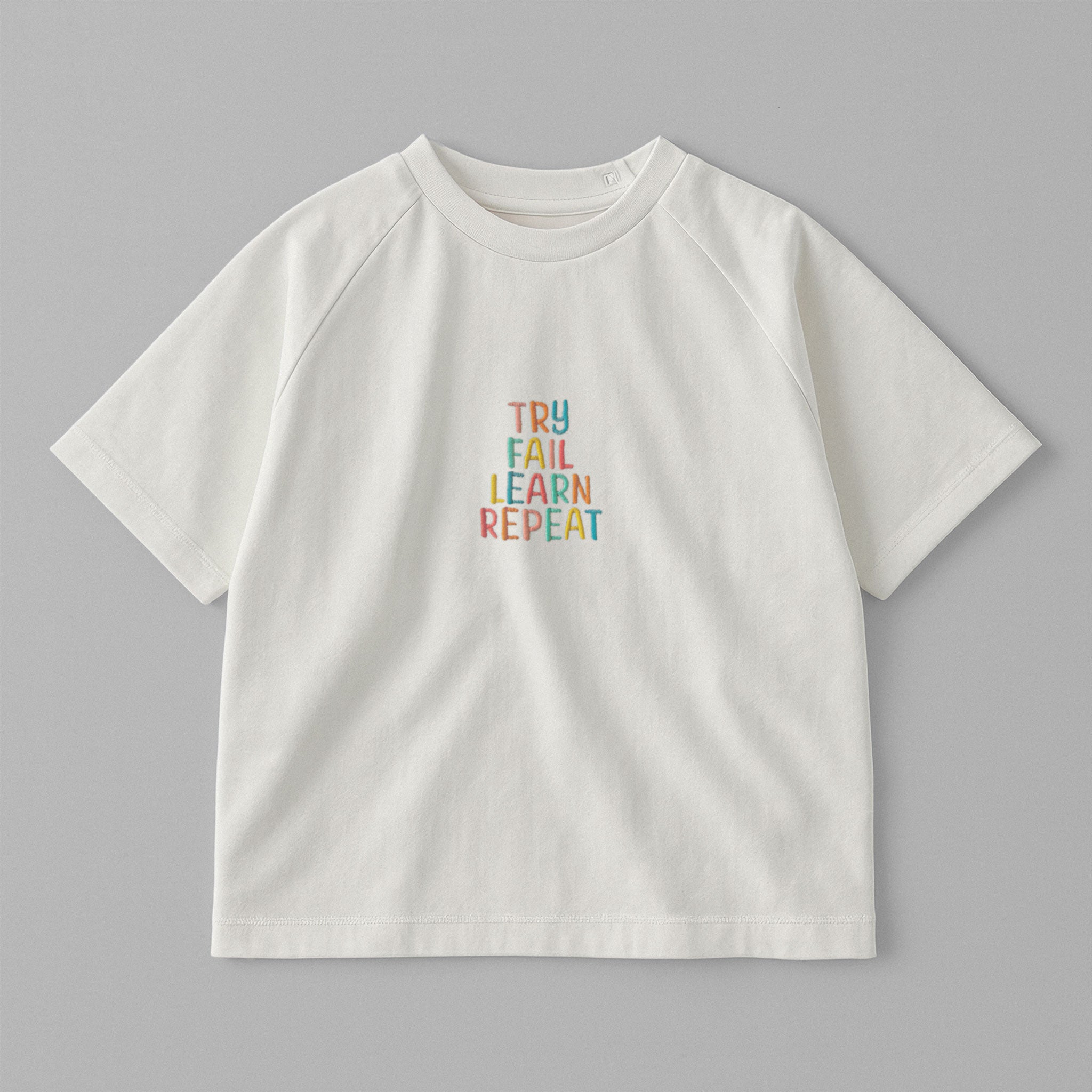 Ekru renkli, reglan kollu, omuzdan çıtçıtlı bebek tişörtü. Göğüs kısmında renkli ipliklerle "TRY FAIL LEARN REPEAT" yazısı işlenmiştir.