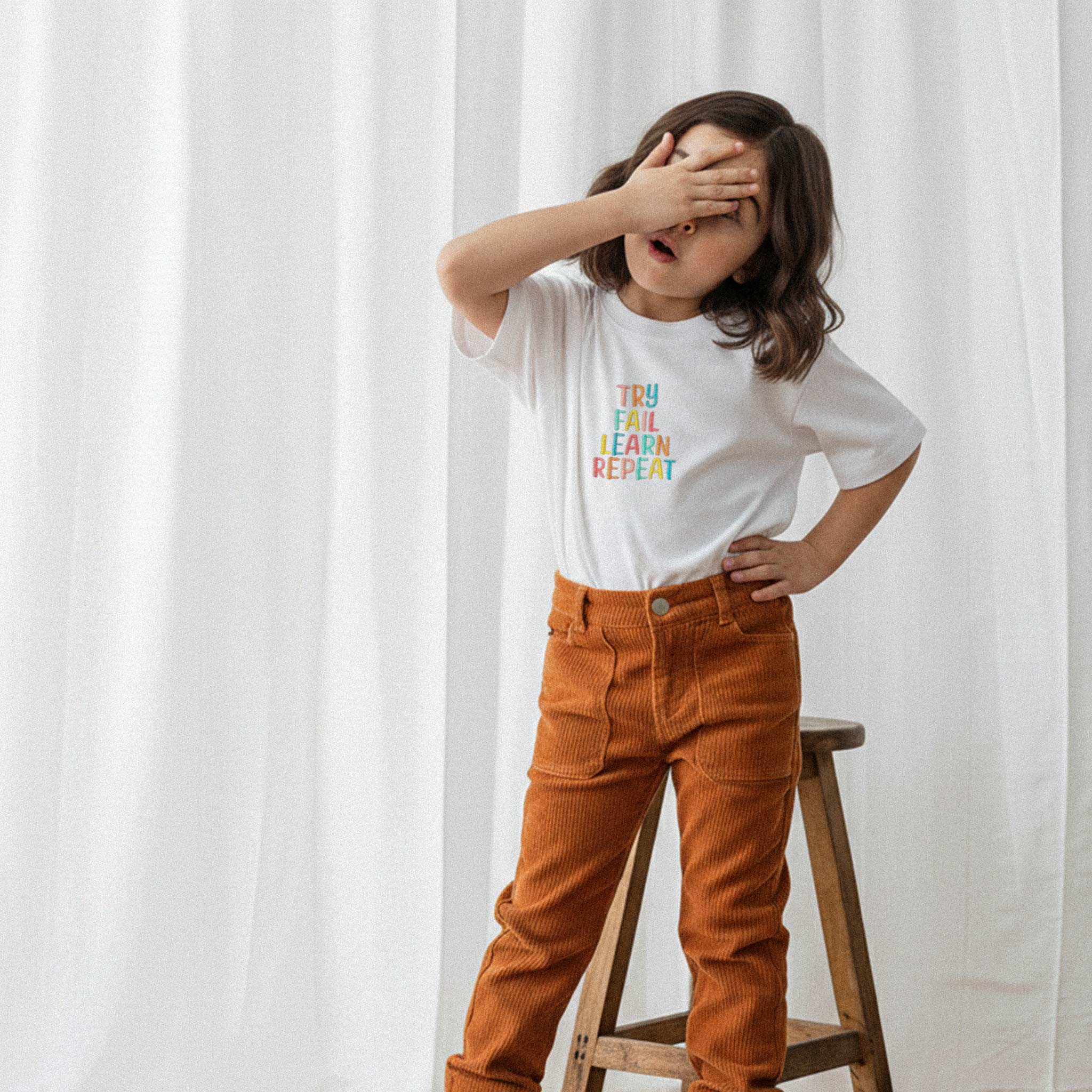 Taba rengi kadife pantolon ve ekru renkli, 'Try Fail Learn Repeat' nakışlı oversize tişört giyen kız çocuk, eliyle gözünü kapatarak havalı bir poz veriyor.