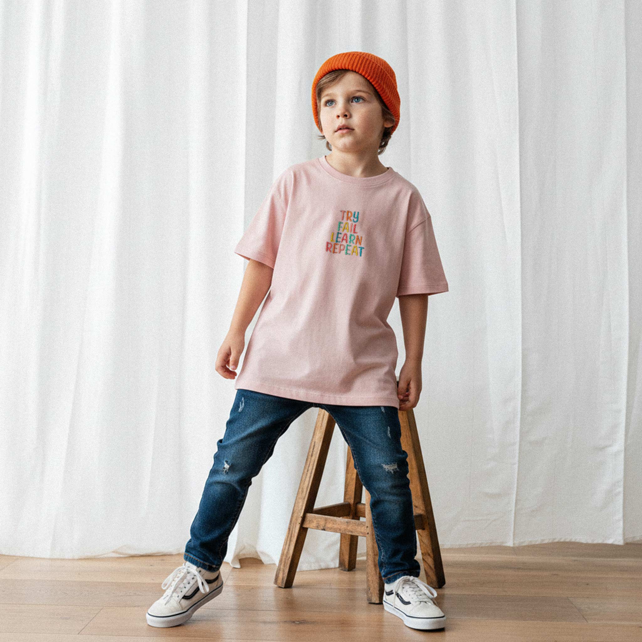 Turuncu bere takan, yırtık jean pantolon ve gül kurusu renkli, 'Try Fail Learn Repeat' nakışlı oversize tişört giyen erkek çocuk, havalı bir şekilde poz veriyor.