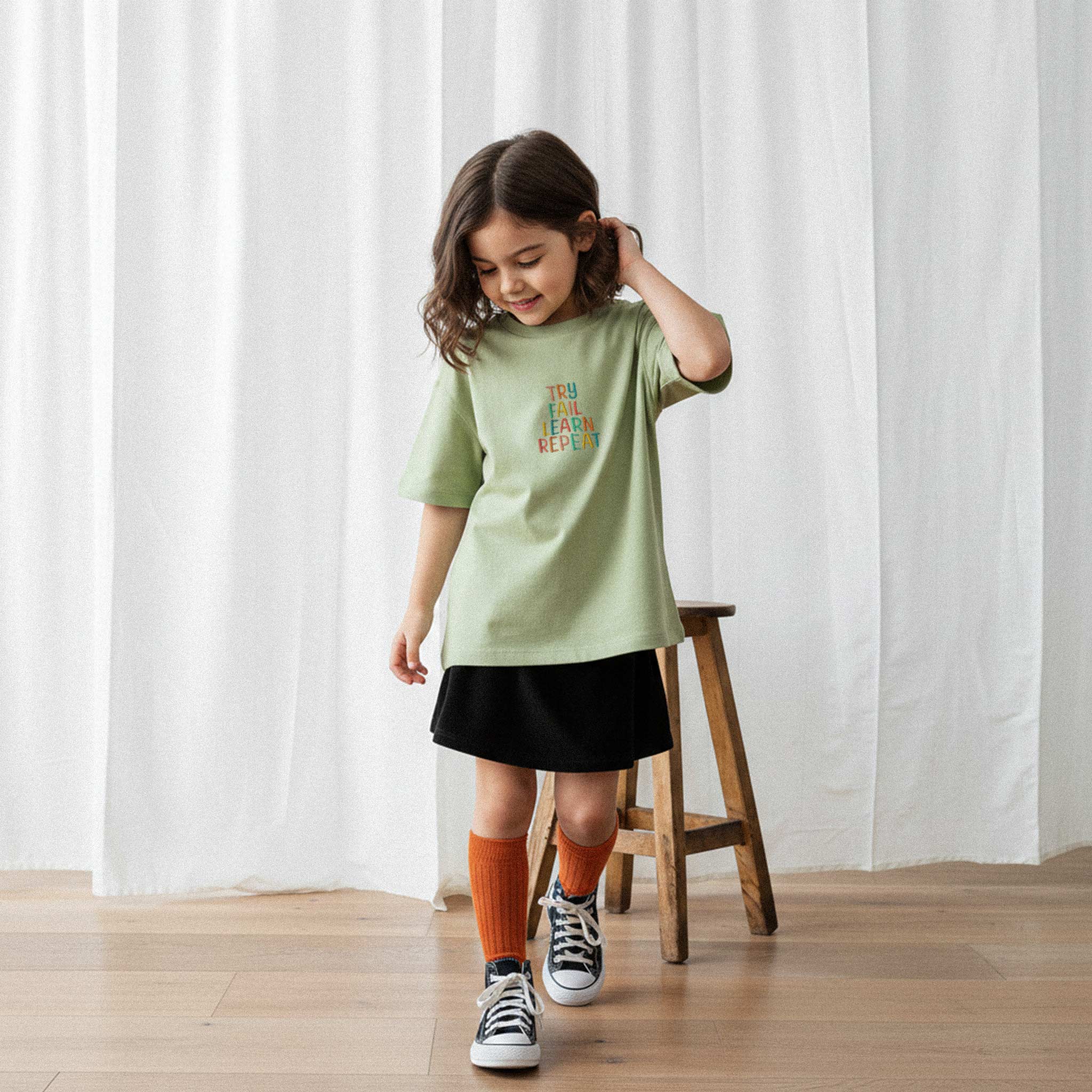 Siyah etek ve haki yeşili, 'Try Fail Learn Repeat' nakışlı oversize tişört giyen kız çocuk, tişörtüne bakarak gülümseyerek yürüyor.