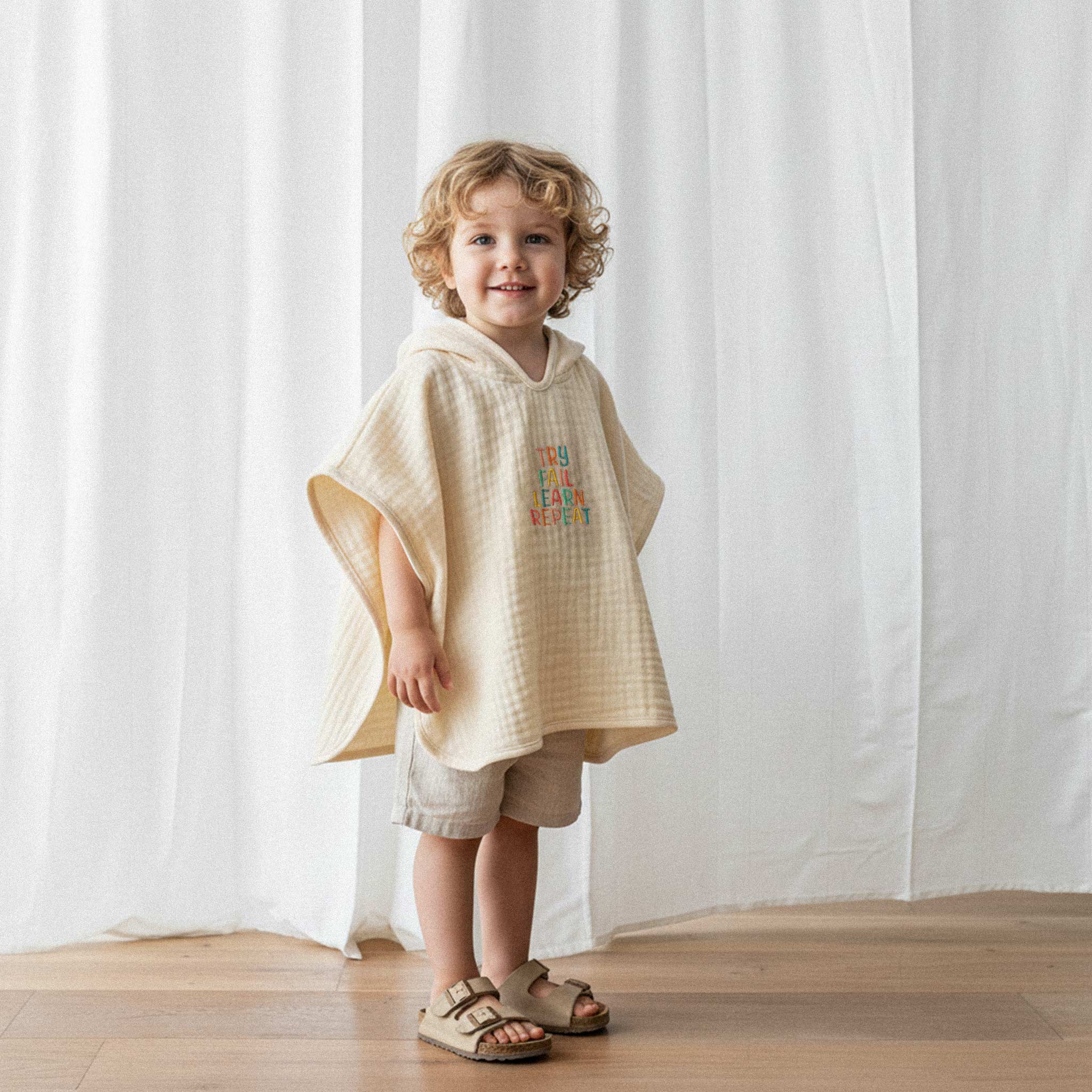 Keten şort ve ekru renkli, 'Try Fail Learn Repeat' nakışlı kapüşonlu müslin panço giyen kıvırcık saçlı küçük erkek çocuk, sevimli bir şekilde gülümseyerek poz veriyor.