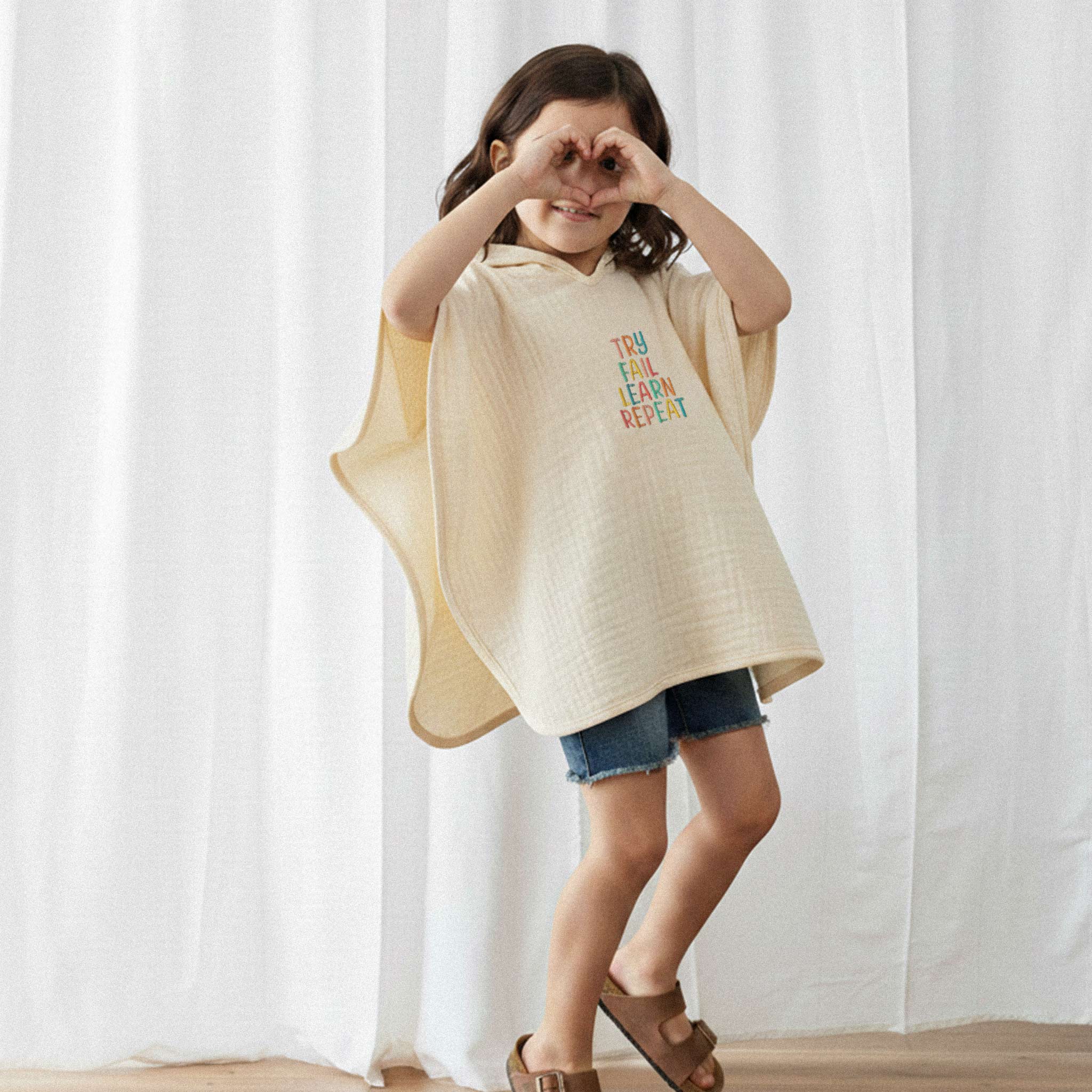Jean şort ve ekru renkli, 'Try Fail Learn Repeat' nakışlı kapüşonlu müslin panço giyen küçük kız çocuk, elleriyle kalp işareti yaparak gülümsüyor.