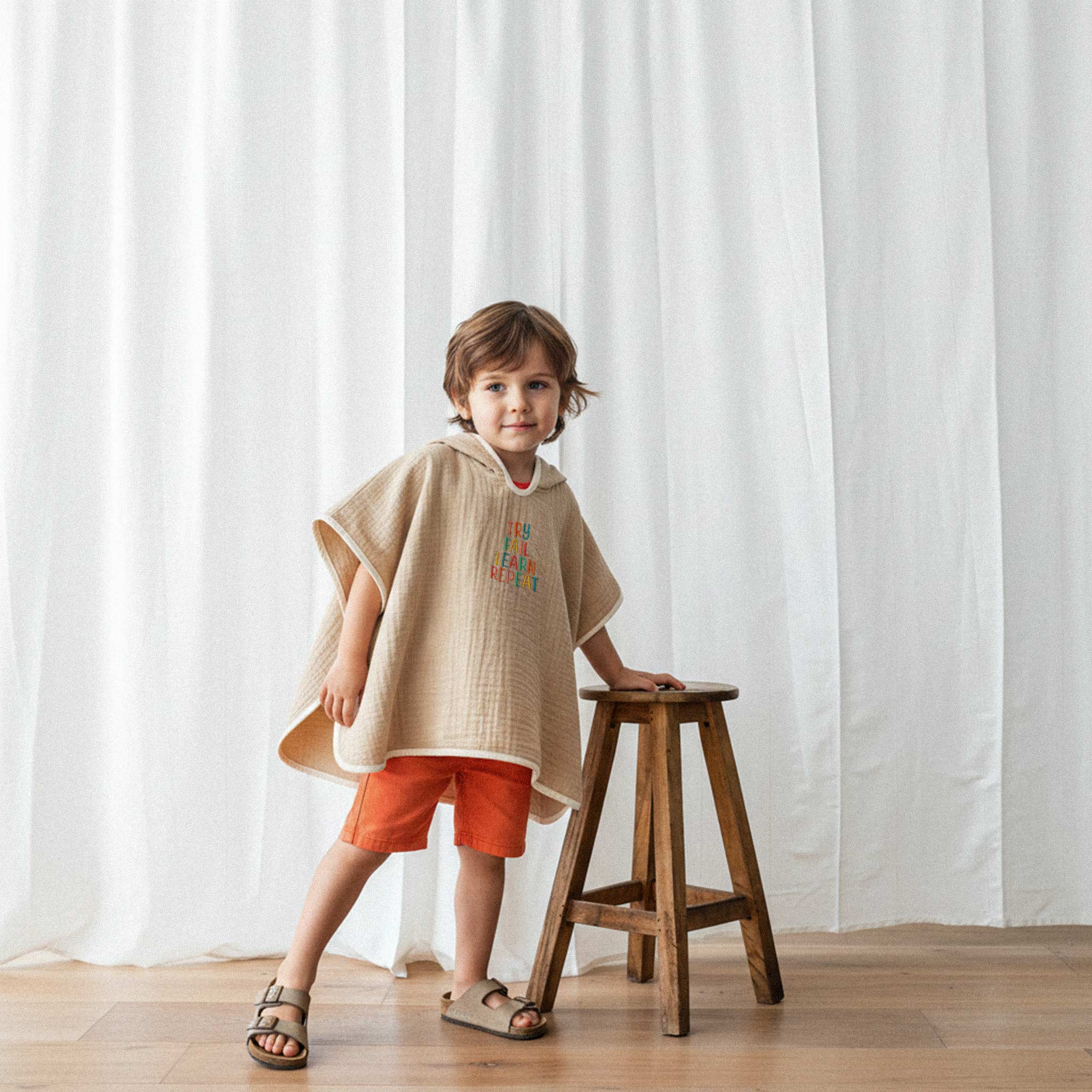 Turuncu şort ve somon rengi, 'Try Fail Learn Repeat' nakışlı kapüşonlu müslin panço giyen erkek çocuk, ahşap bir tabureye yaslanarak poz veriyor.