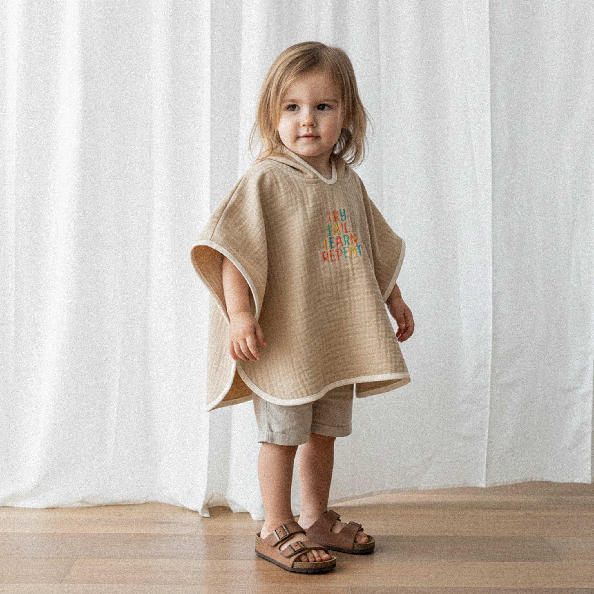 Bej şort ve somon rengi, 'Try Fail Learn Repeat' nakışlı kapüşonlu müslin panço giyen küçük kız çocuk, sevimli bir şekilde poz veriyor.