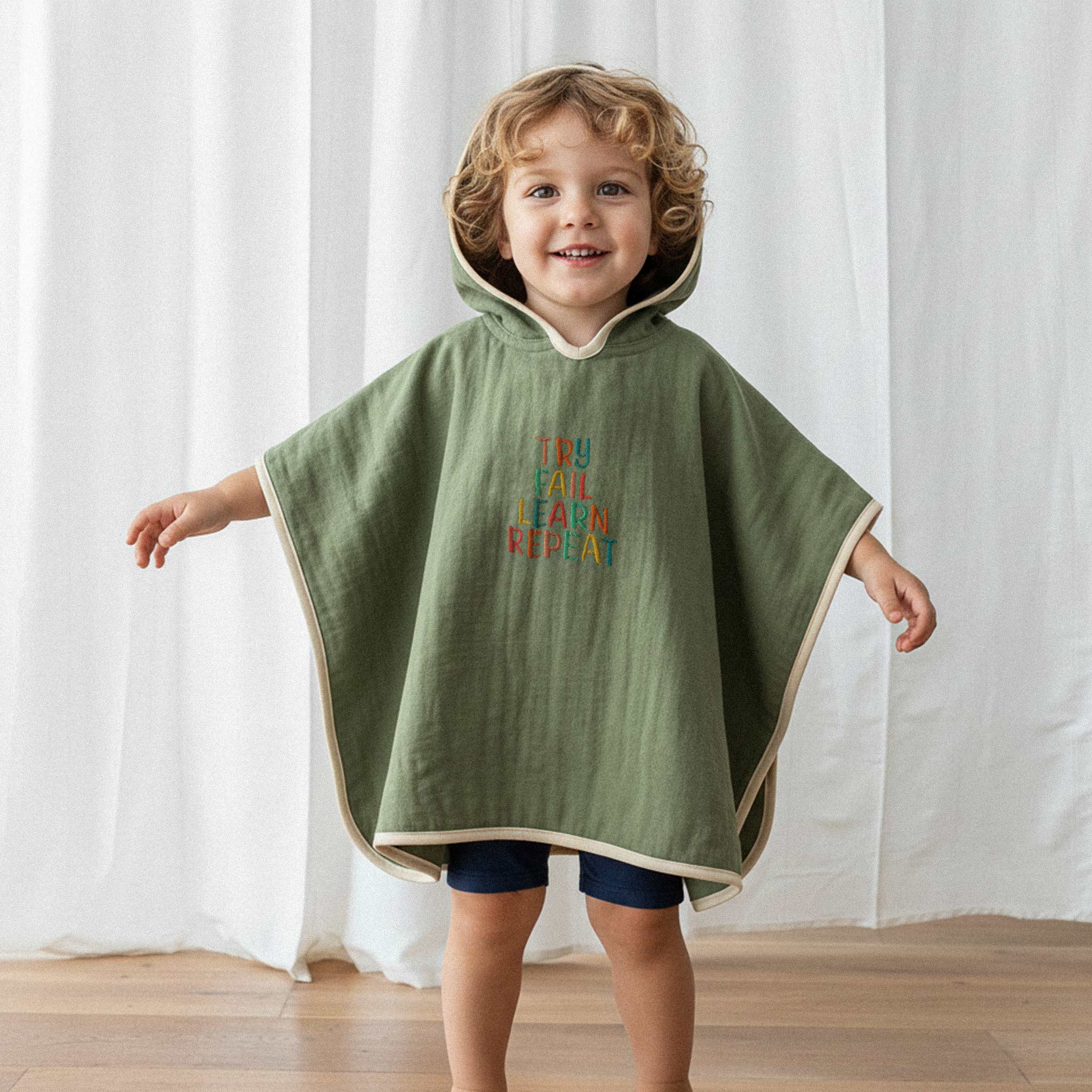 Lacivert şort ve haki yeşili, 'Try Fail Learn Repeat' nakışlı kapüşonlu müslin panço giyen kıvırcık saçlı küçük erkek çocuk, kollarını açarak neşeyle gülümsüyor.