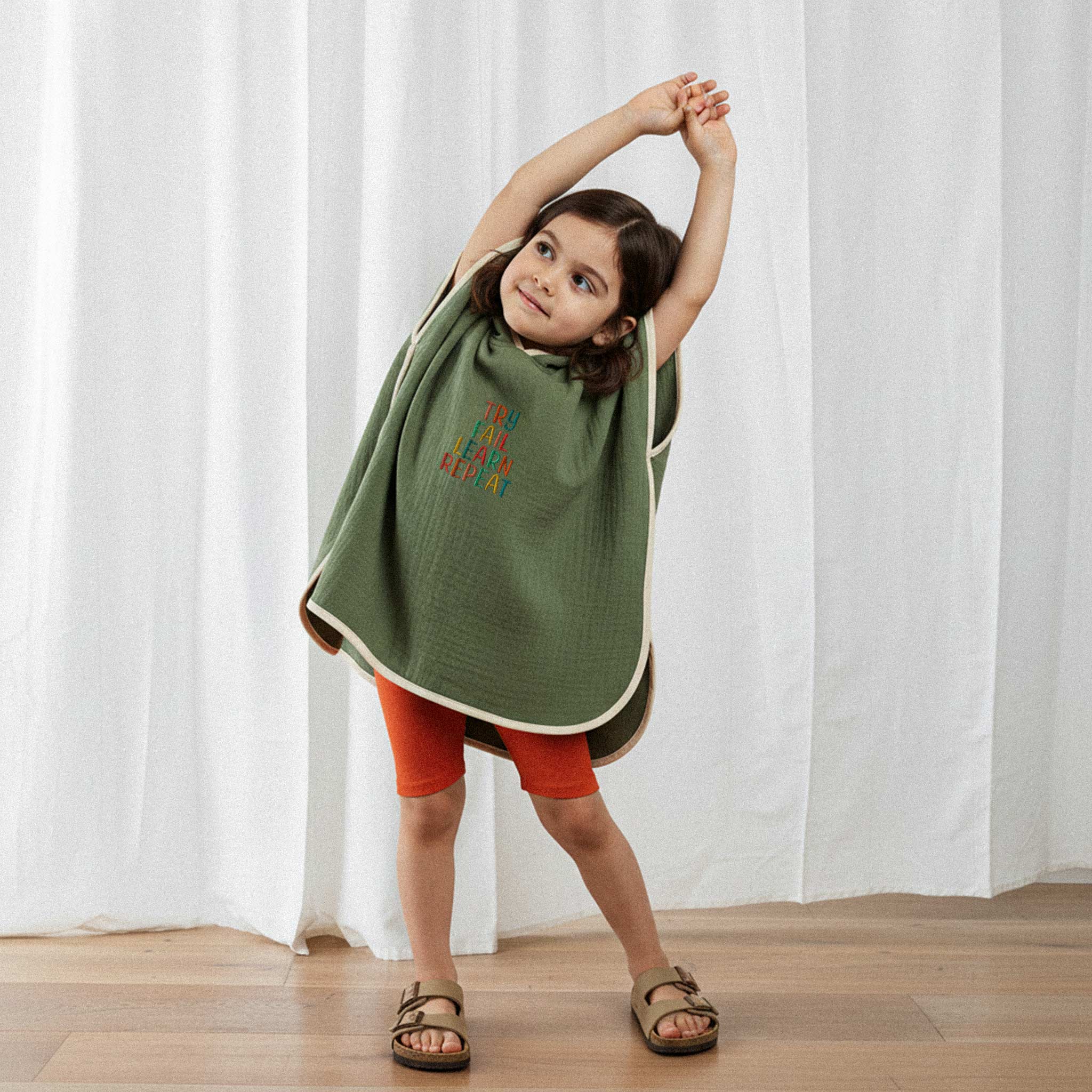 Turuncu şort ve haki yeşili, 'Try Fail Learn Repeat' nakışlı kapüşonlu müslin panço giyen kız çocuk, kollarını yukarı kaldırarak esneme hareketi yapıyor.