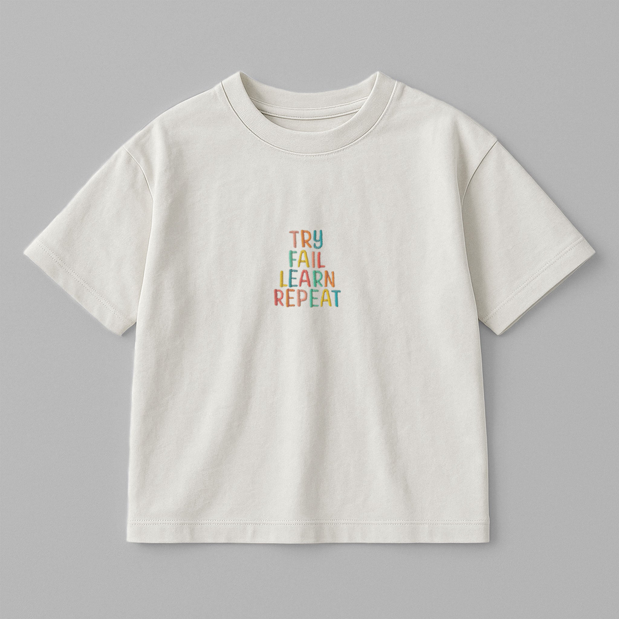 Ekru renkli, oversize kesim, 1-5 yaş için pamuklu çocuk tişörtü. Göğüs kısmında renkli ipliklerle "TRY FAIL LEARN REPEAT" yazısı işlenmiştir.