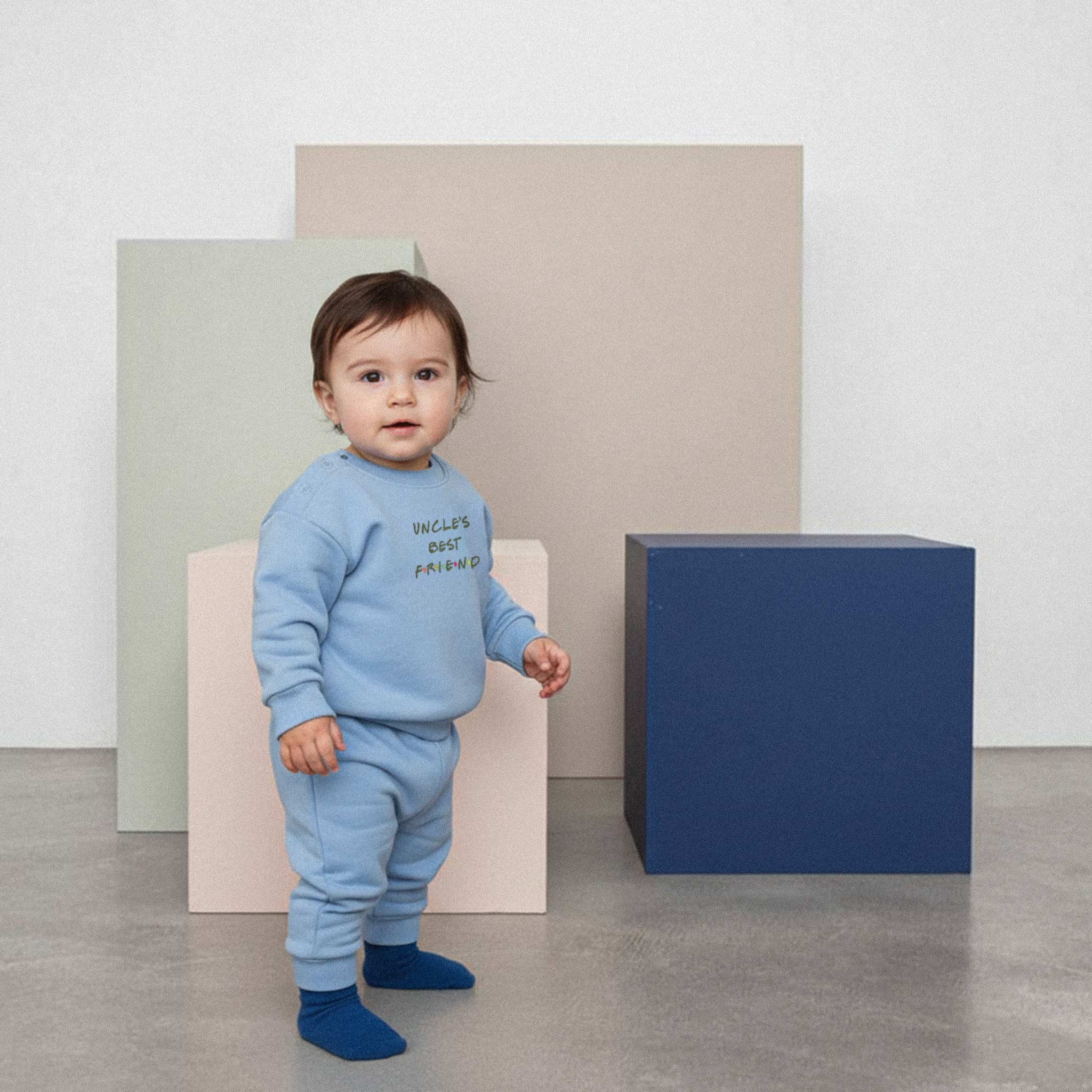 Üzerinde mavi renkli, 'Uncle's Best Friend' nakışlı eşofman takımı bulunan sevimli bebek, renkli küplerin önünde ayakta durarak kameraya bakıyor.
