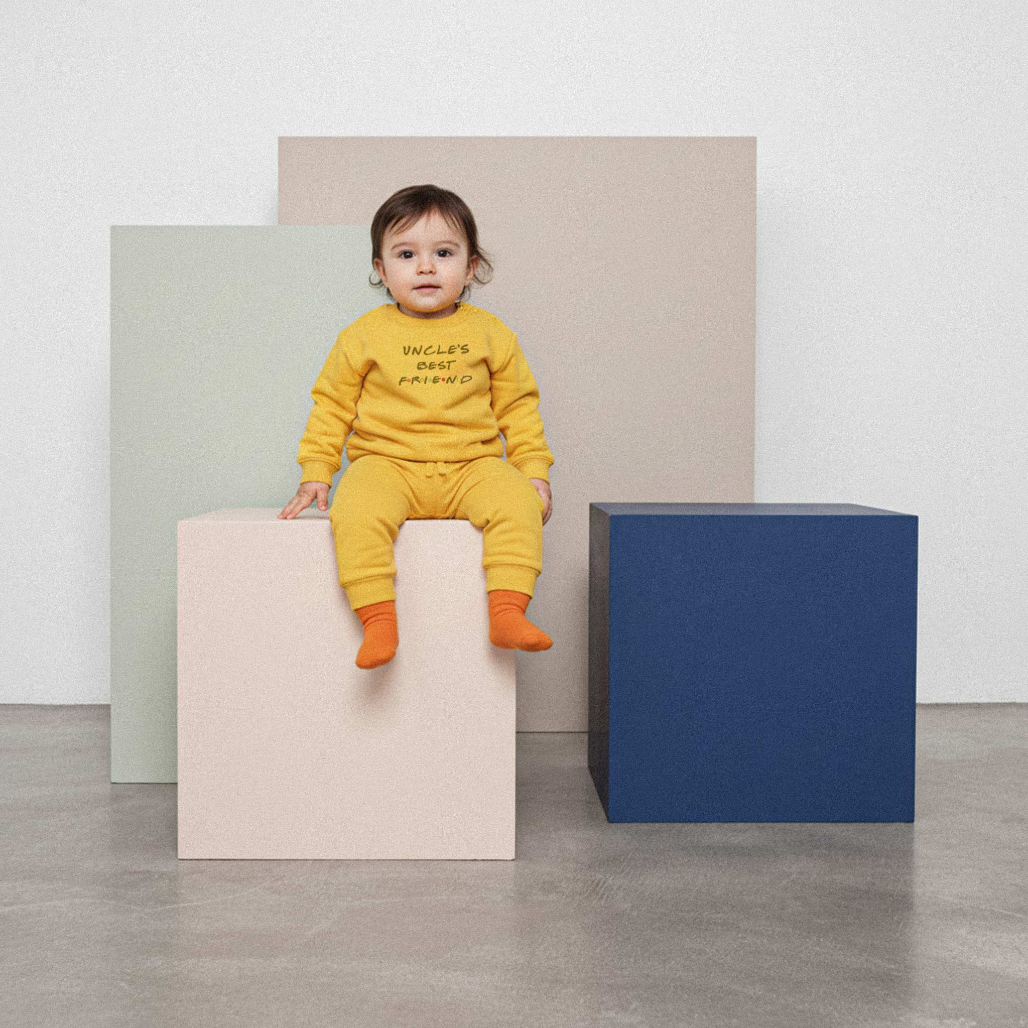 Üzerinde hardal sarısı renkli, 'Uncle's Best Friend' nakışlı eşofman takımı bulunan sevimli bebek, renkli bir küpün üzerinde oturarak kameraya bakıyor.