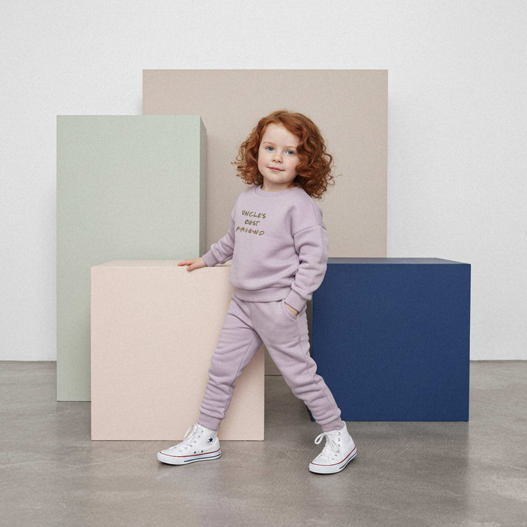 Kızıl saçlı, lila renkli 'Uncle's Best Friend' eşofman takımı giyen kız çocuk, bir eli cebinde renkli bir küpe yaslanarak poz veriyor.