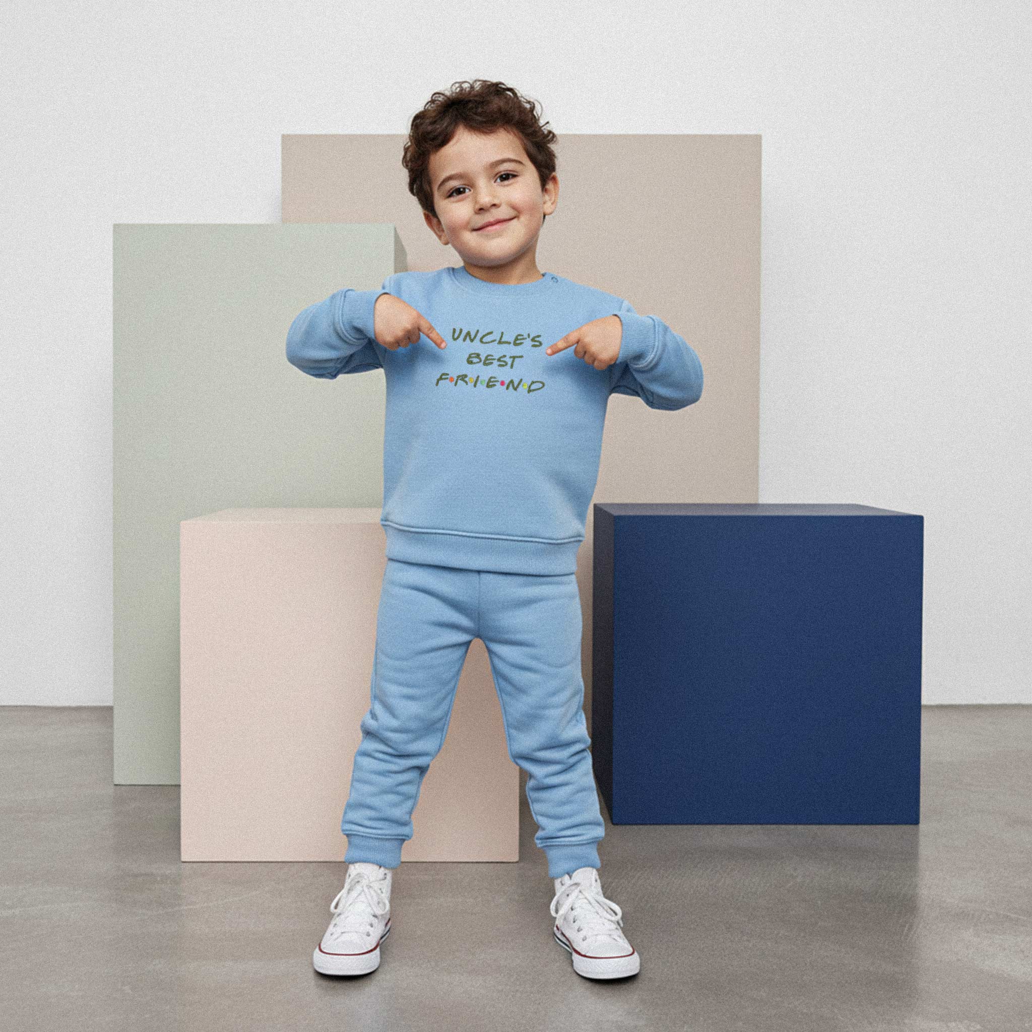Kıvırcık saçlı, mavi renkli 'Uncle's Best Friend' eşofman takımı giyen erkek çocuk, gülümseyerek sweatshirt üzerindeki nakışı işaret ediyor.