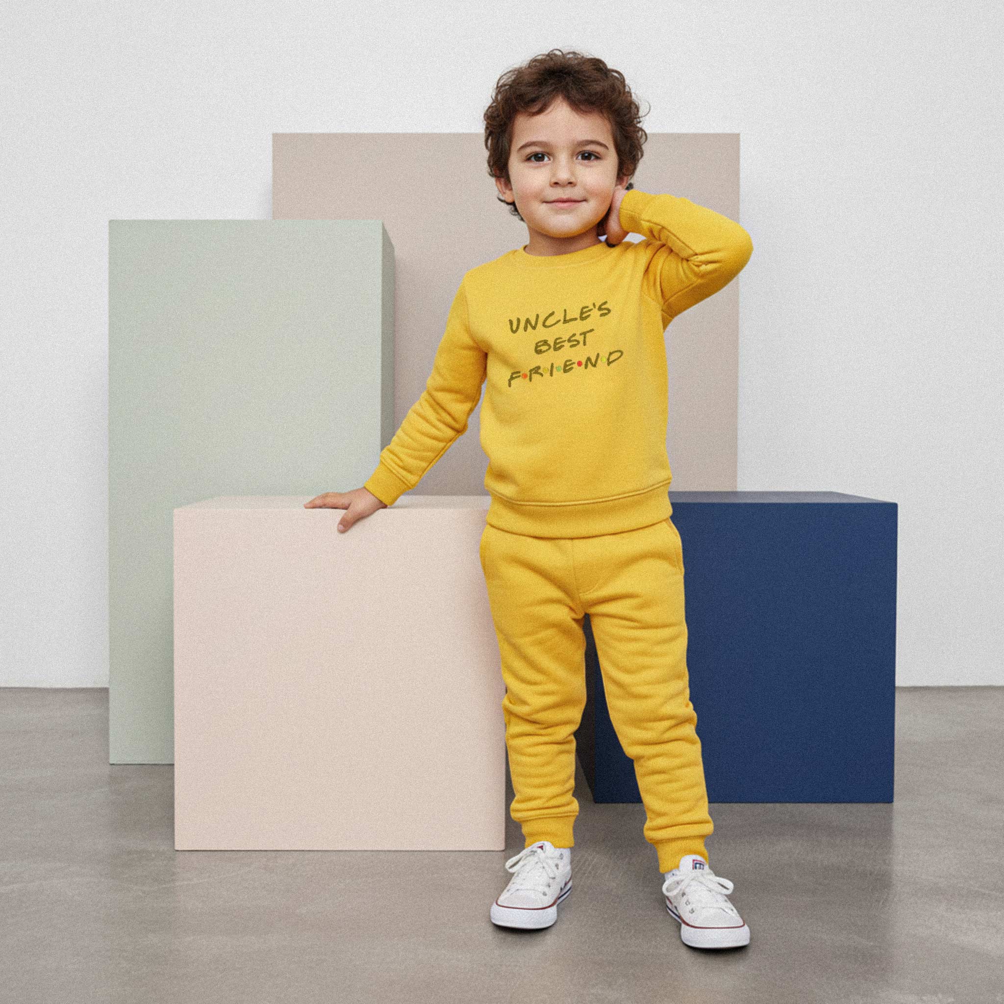 Kıvırcık saçlı, hardal sarısı renkli 'Uncle's Best Friend' eşofman takımı giyen erkek çocuk, renkli küplerin önünde durarak gülümseyerek poz veriyor.