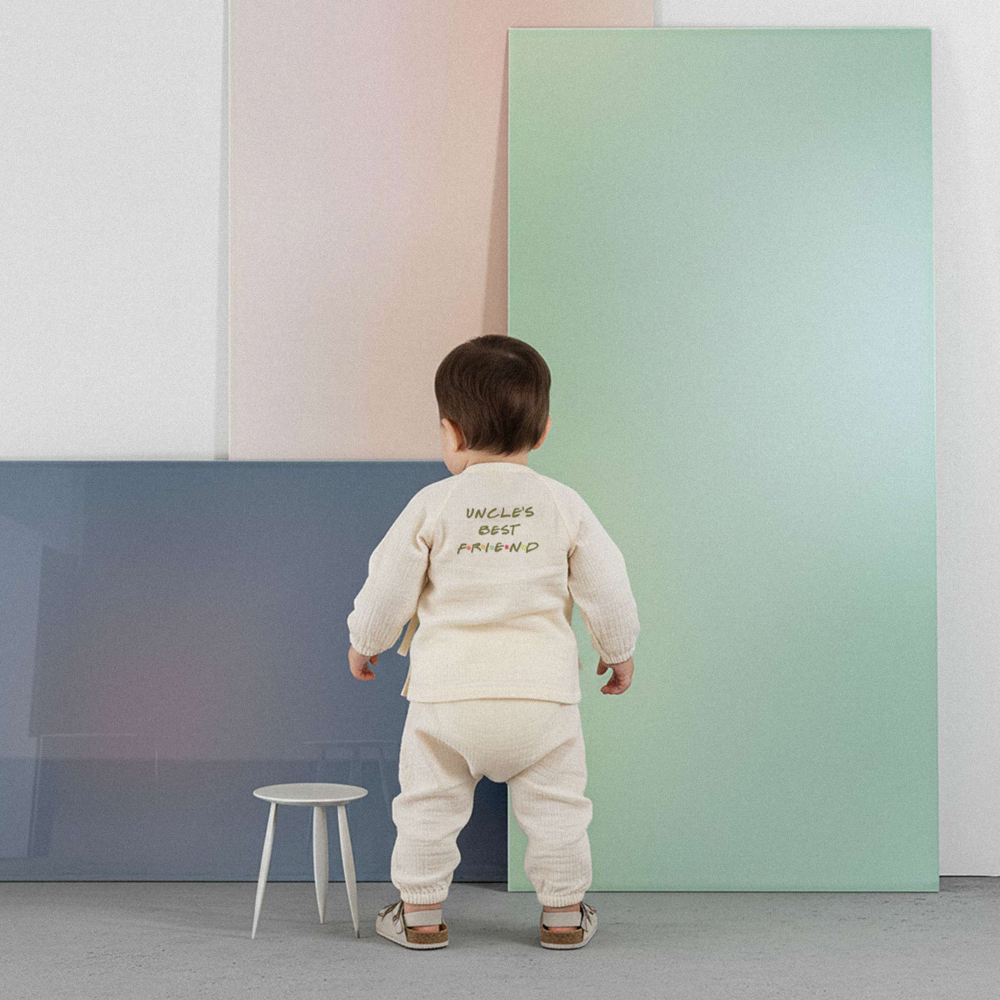 Üzerinde ekru renkli, 'Uncle's Best Friend' baskılı müslin kimono takımı bulunan sevimli bebek, renkli küplerin önünde ayakta durarak poz veriyor.