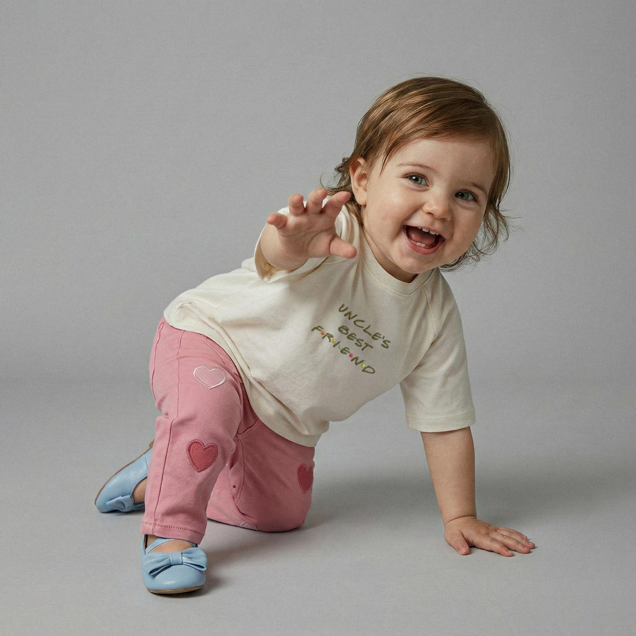 Ekru renkli 'Uncle's Best Friend' nakışlı tişört ve kalpli pembe pantolon giyerek elini kameraya doğru uzatan ve neşeyle gülen kız bebek model.