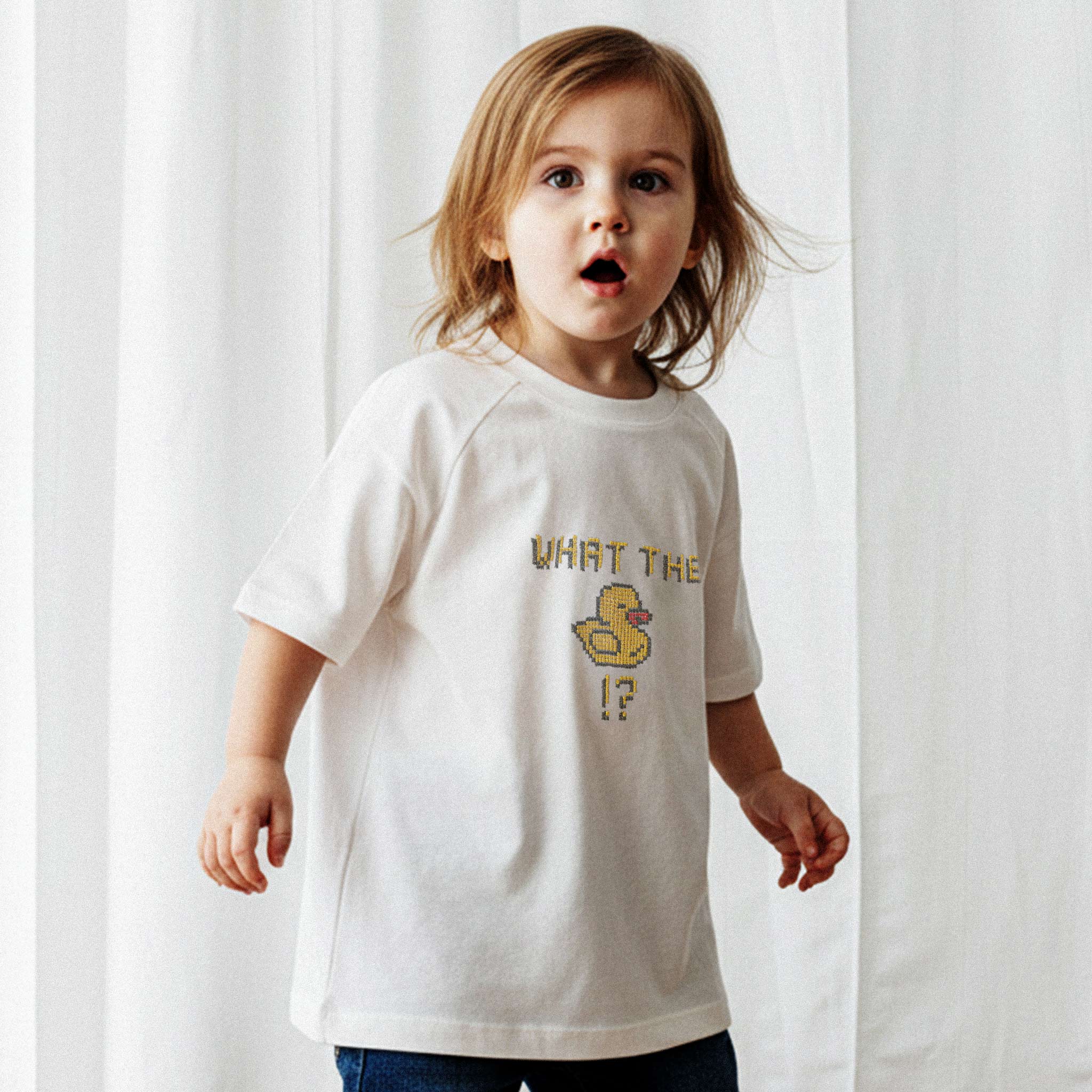 Üzerinde ekru renkli, 'What The Duck!?' nakışlı reglan kollu oversize bebek tişörtü giyen küçük kız çocuk, şaşkın ve sevimli bir ifadeyle kameraya bakıyor.