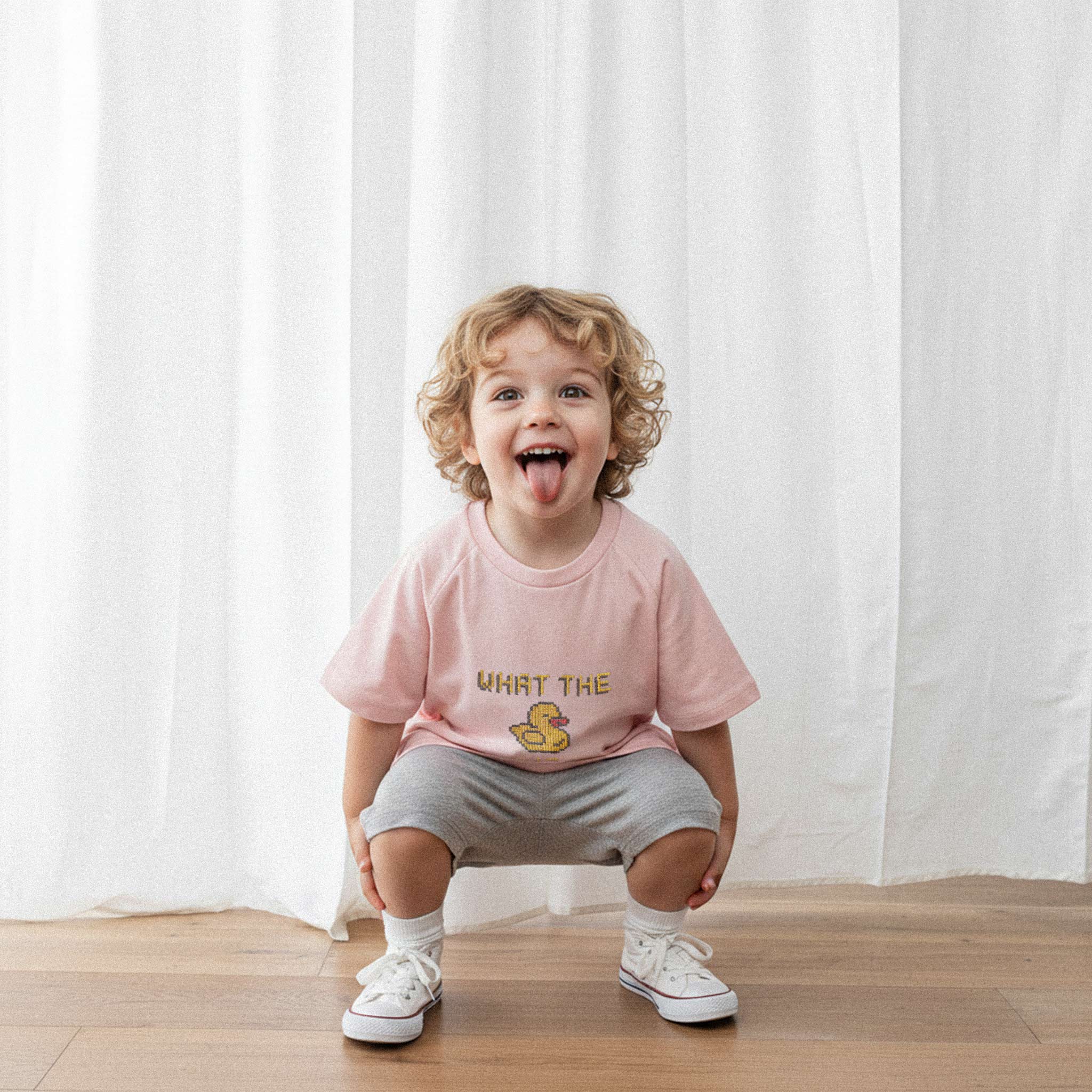 Gri şort ve gül kurusu renkli, 'What The Duck!?' nakışlı reglan kollu oversize bebek tişörtü giyen kıvırcık saçlı küçük erkek çocuk, çömelmiş ve dilini çıkararak neşeyle poz veriyor.