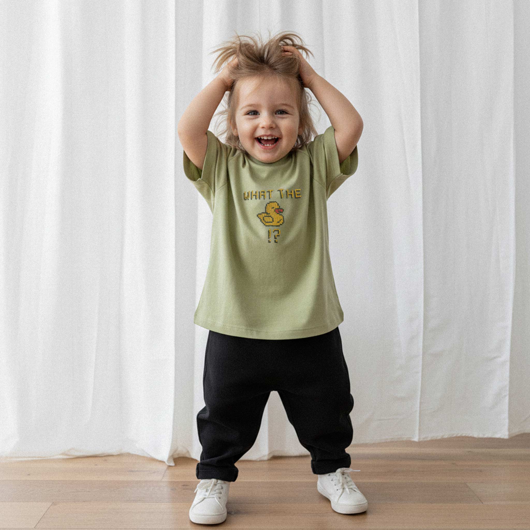 Üzerinde haki yeşili renkli, 'What The Duck!?' nakışlı reglan kollu oversize bebek tişörtü giyen küçük kız çocuk, elleriyle saçlarını karıştırarak neşeyle kahkaha atıyor.