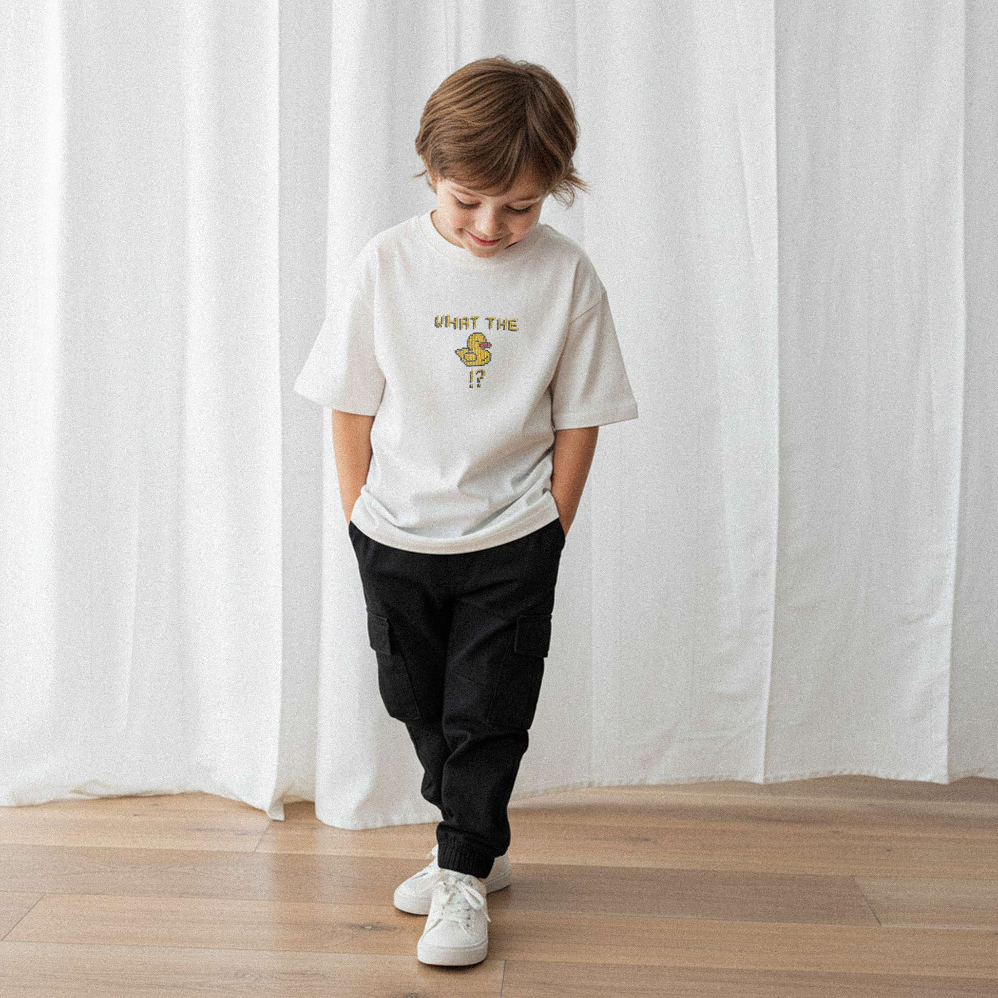 Siyah kargo pantolon ve ekru renkli, 'What The Duck!?' nakışlı oversize tişört giyen erkek çocuk, elleri ceplerinde tişörtüne bakarak gülümsüyor.