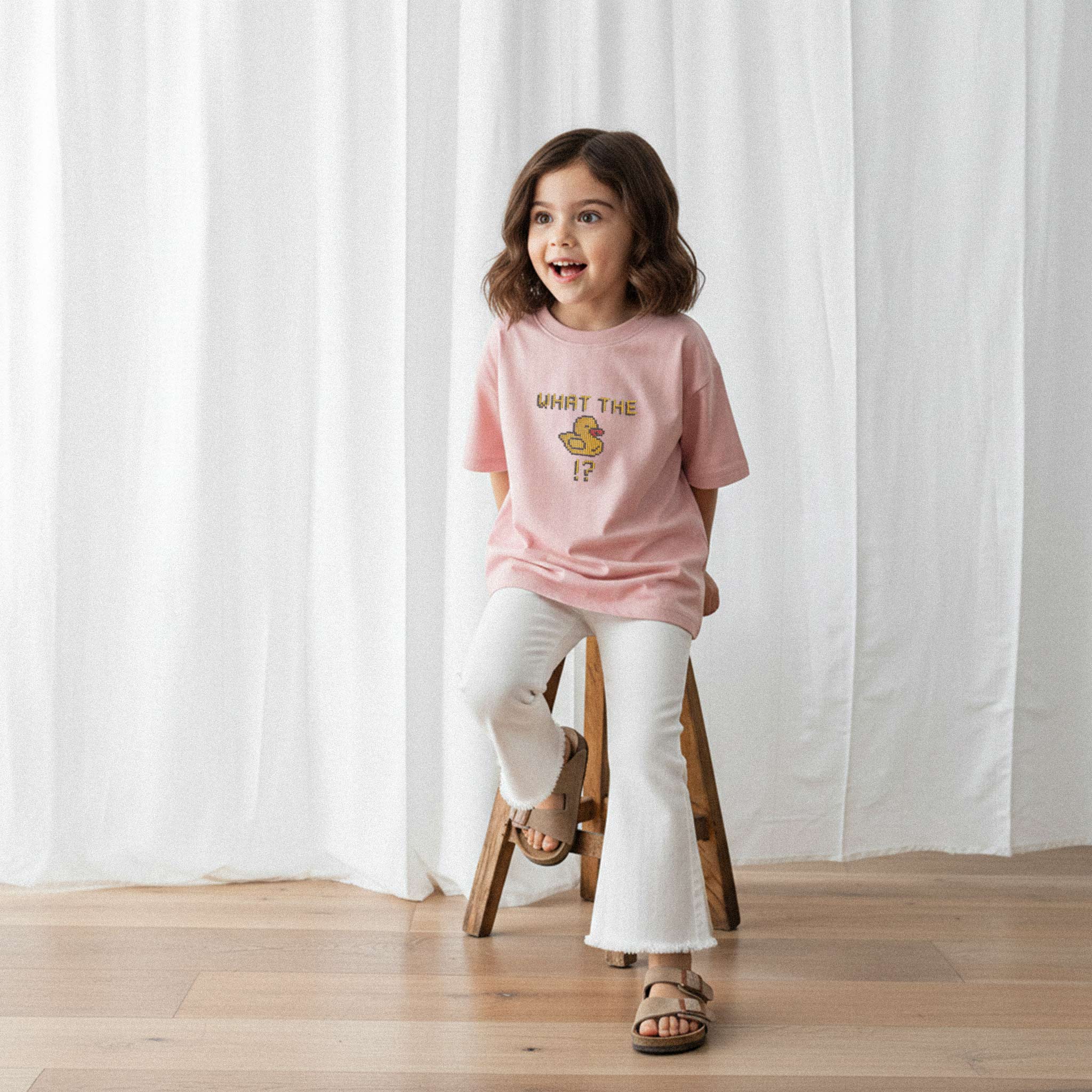 Beyaz İspanyol paça pantolon ve gül kurusu, 'What The Duck!?' nakışlı oversize tişört giyen kız çocuk, sandalyede otururken neşeyle yana bakıyor.