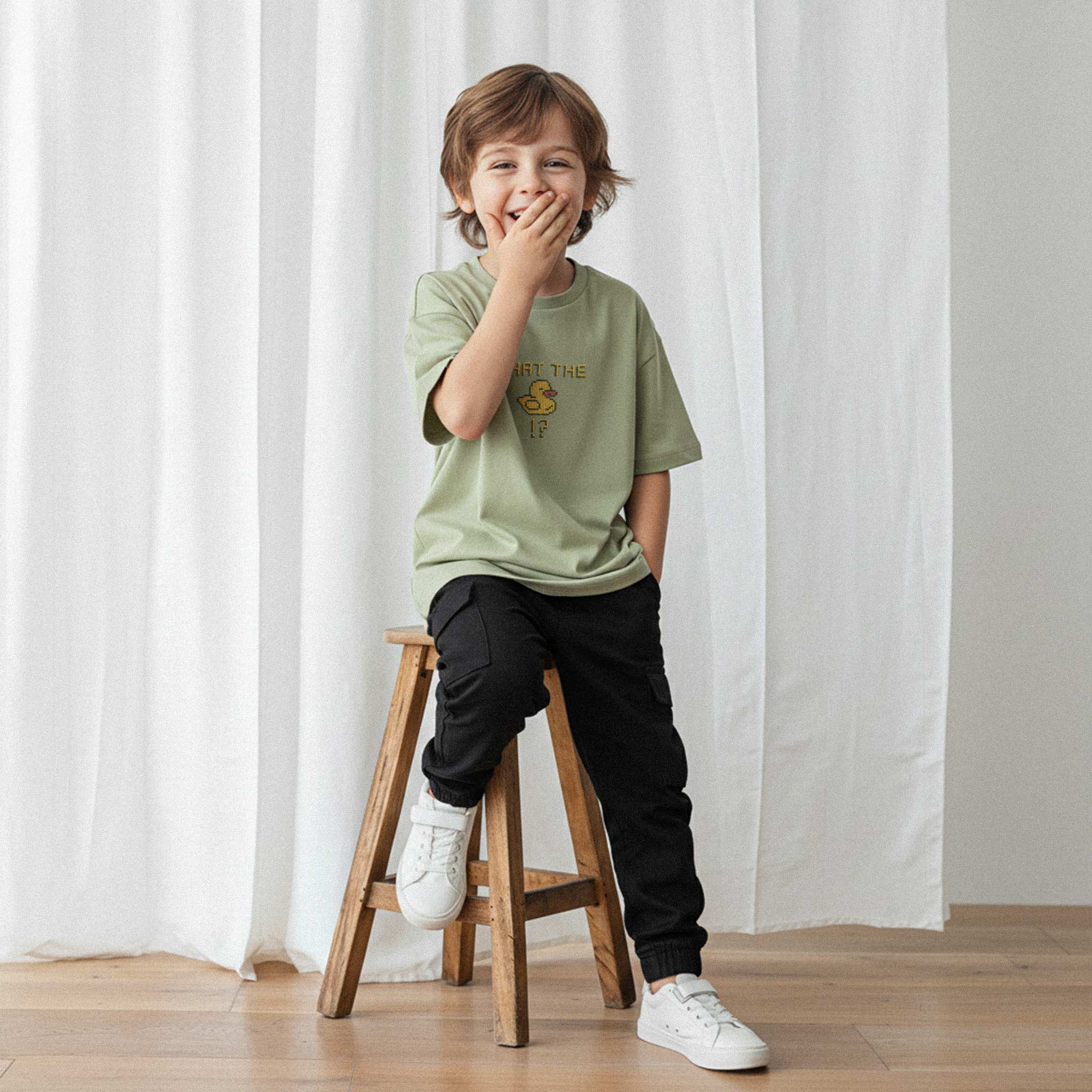 Siyah kargo pantolon ve haki yeşili, 'What The Duck!?' nakışlı oversize tişört giyen erkek çocuk, sandalyede otururken eliyle ağzını kapatarak gülüyor.