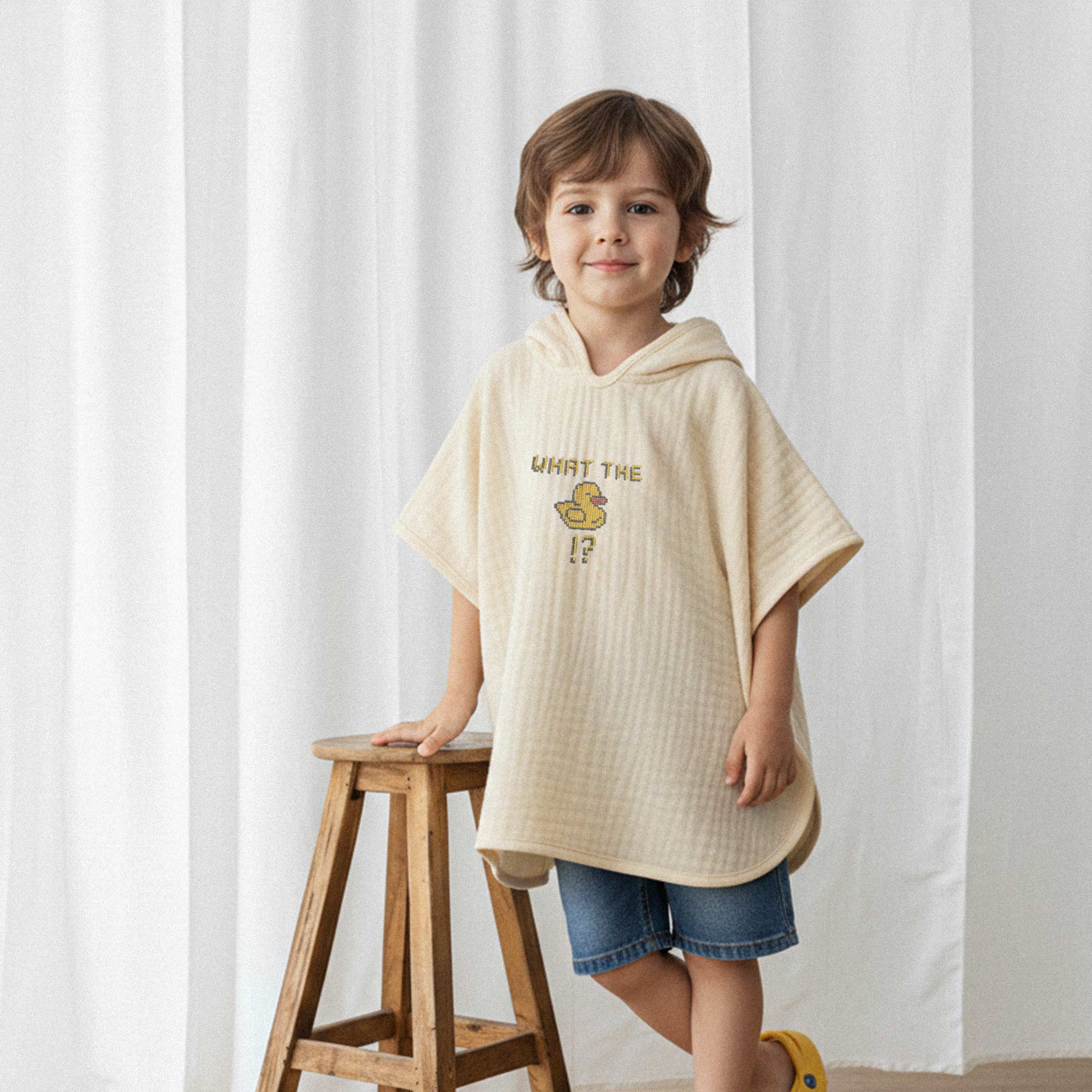 Jean şort ve ekru renkli, 'What The Duck!?' nakışlı kapüşonlu müslin panço giyen erkek çocuk, ahşap bir tabureye yaslanarak gülümsüyor.