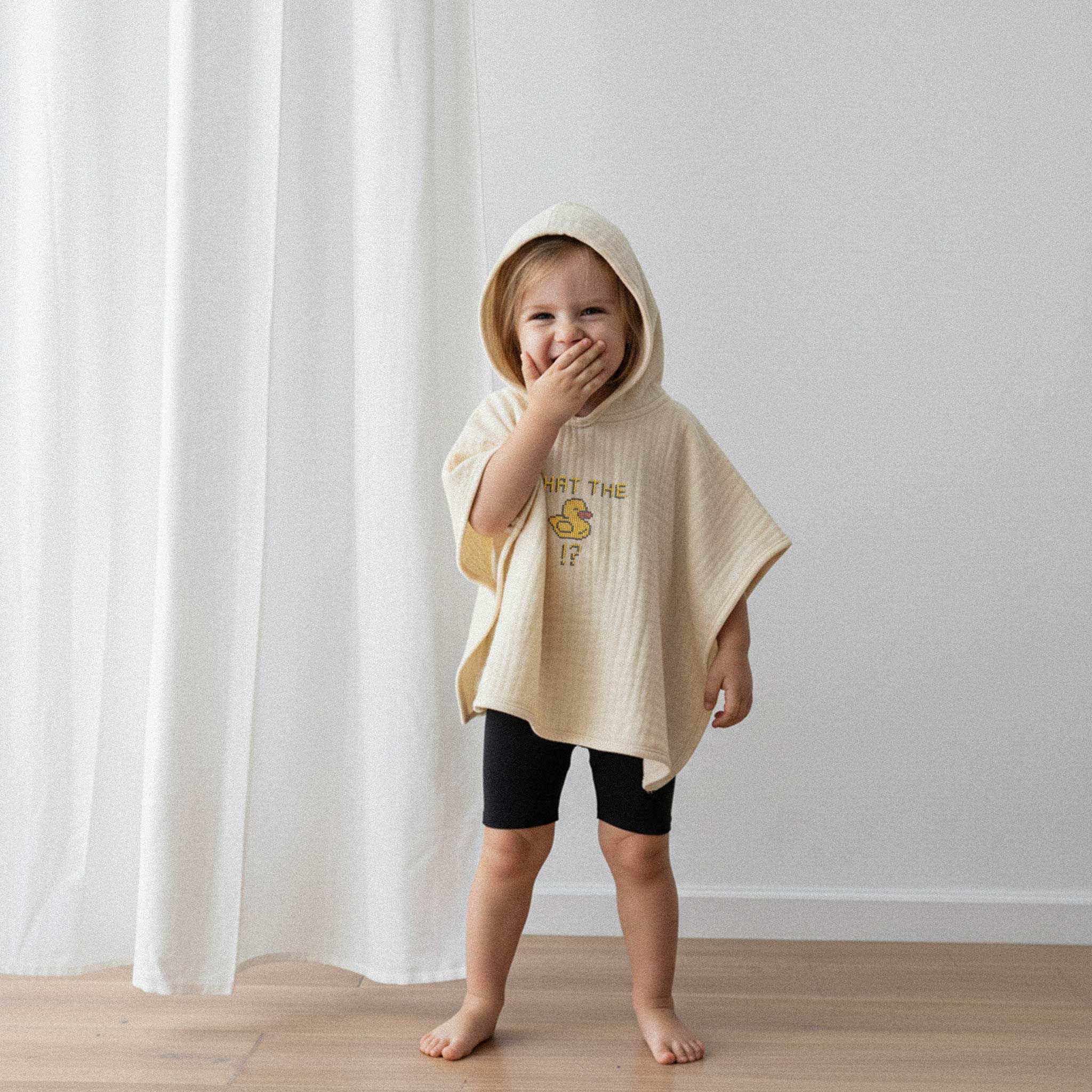 Siyah şort ve ekru renkli, 'What The Duck!?' nakışlı kapüşonlu müslin panço giyen küçük kız çocuk, eliyle ağzını kapatarak kıkırdıyor.
