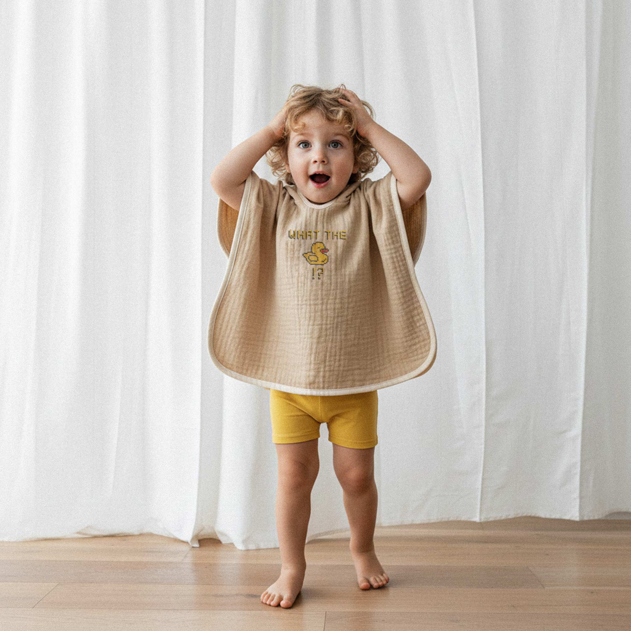 Sarı şort ve somon rengi, 'What The Duck!?' nakışlı kapüşonlu müslin panço giyen kıvırcık saçlı küçük erkek çocuk, elleri başında şaşkın bir surat ifadesiyle poz veriyor.