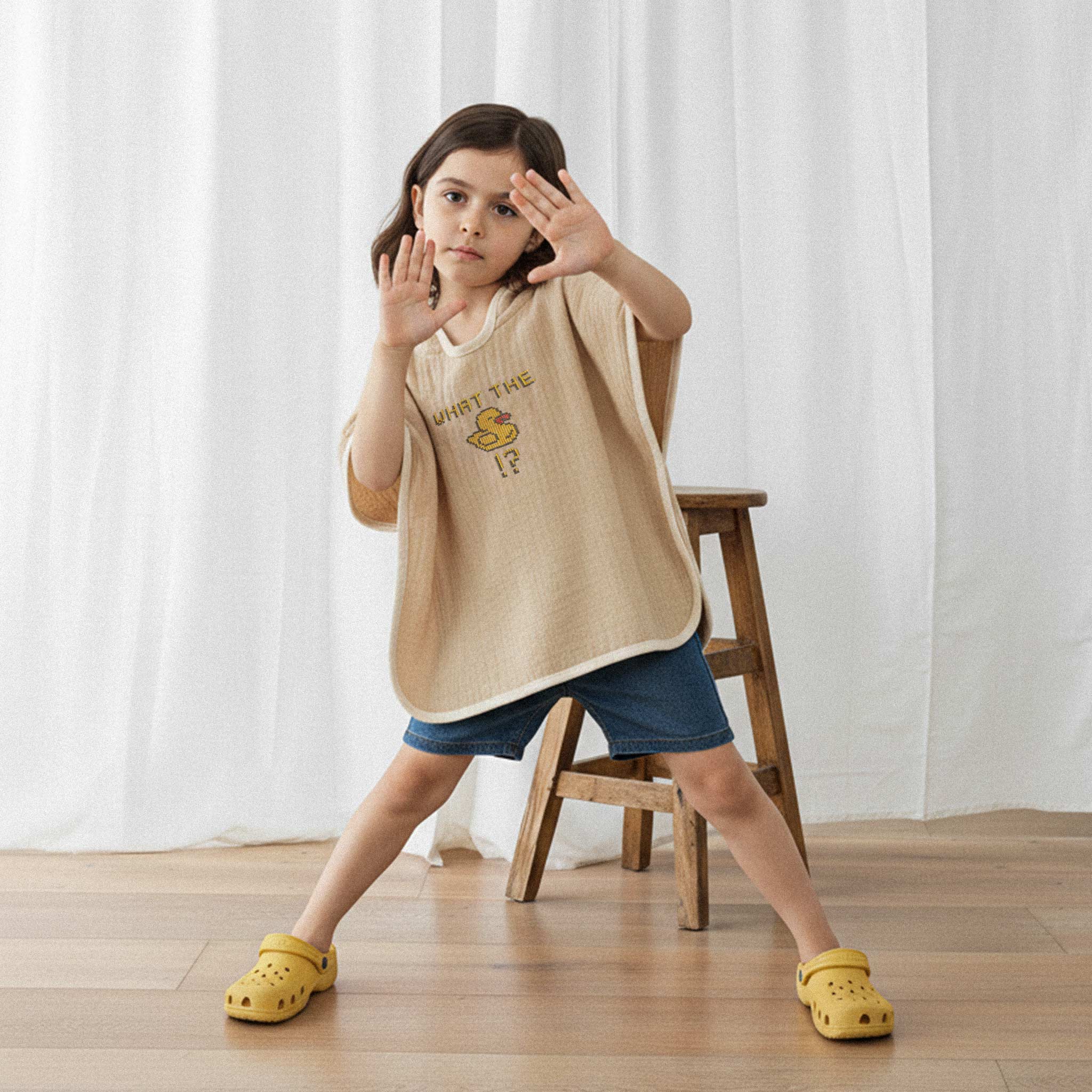 Jean şort ve sarı terlik giyen kız çocuk, üzerinde somon rengi, 'What The Duck!?' nakışlı kapüşonlu müslin panço ile ellerini kaldırarak poz veriyor.
