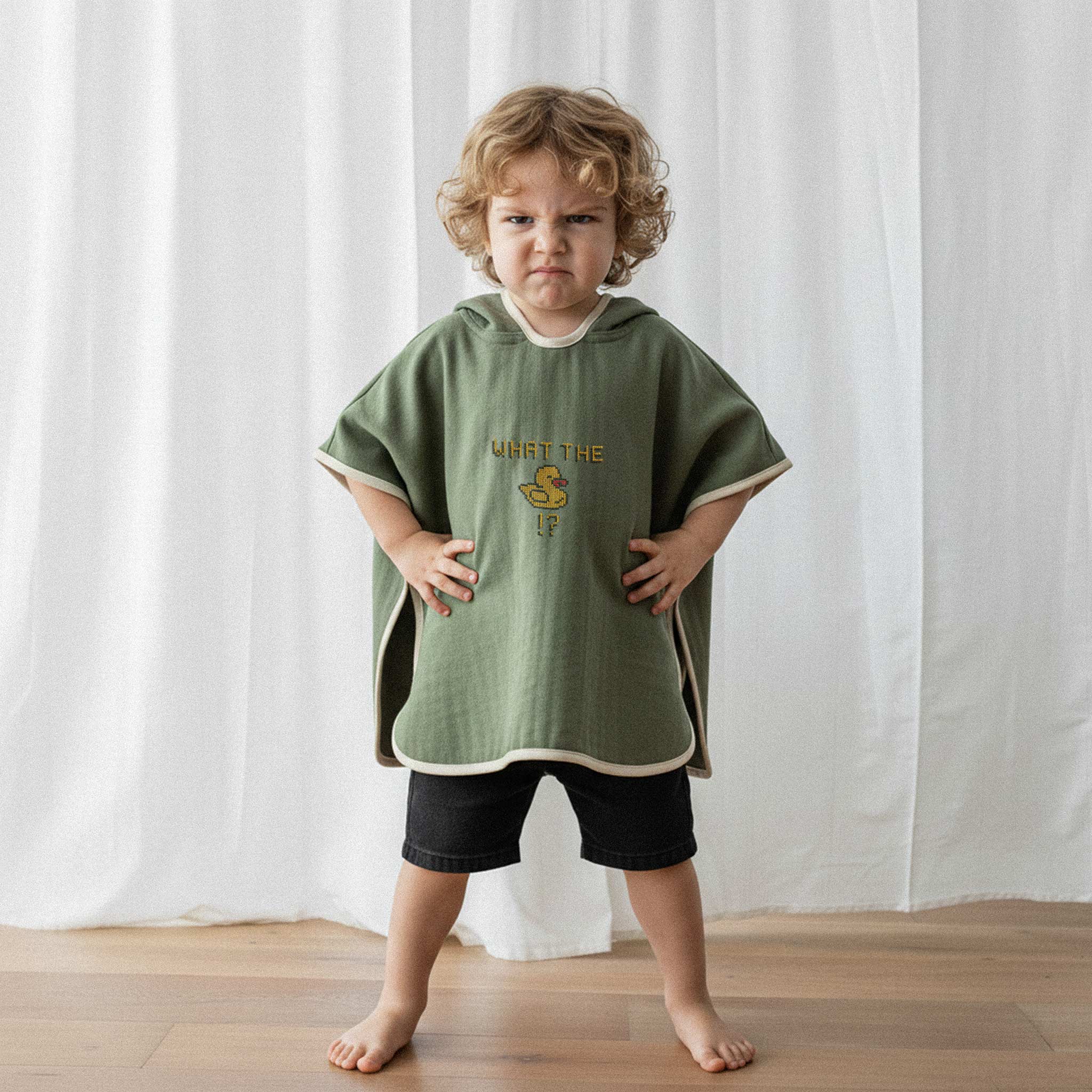 Siyah şort ve haki yeşili, 'What The Duck!?' nakışlı kapüşonlu müslin panço giyen kıvırcık saçlı küçük erkek çocuk, elleri belinde kızgın bir surat ifadesiyle poz veriyor.