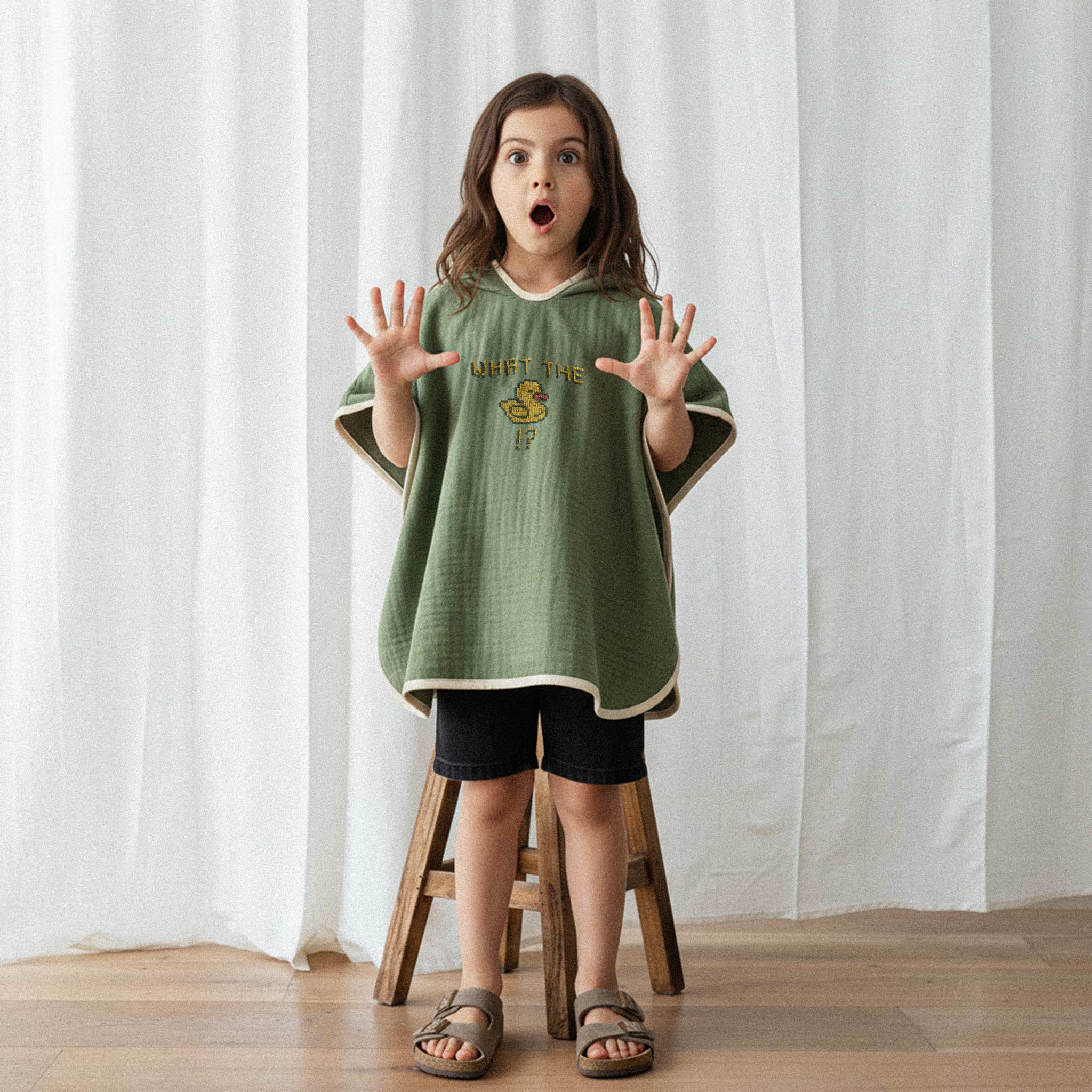 Siyah şort ve haki yeşili, 'What The Duck!?' nakışlı kapüşonlu müslin panço giyen küçük kız çocuk, ellerini kaldırarak şaşkın bir yüz ifadesiyle poz veriyor.